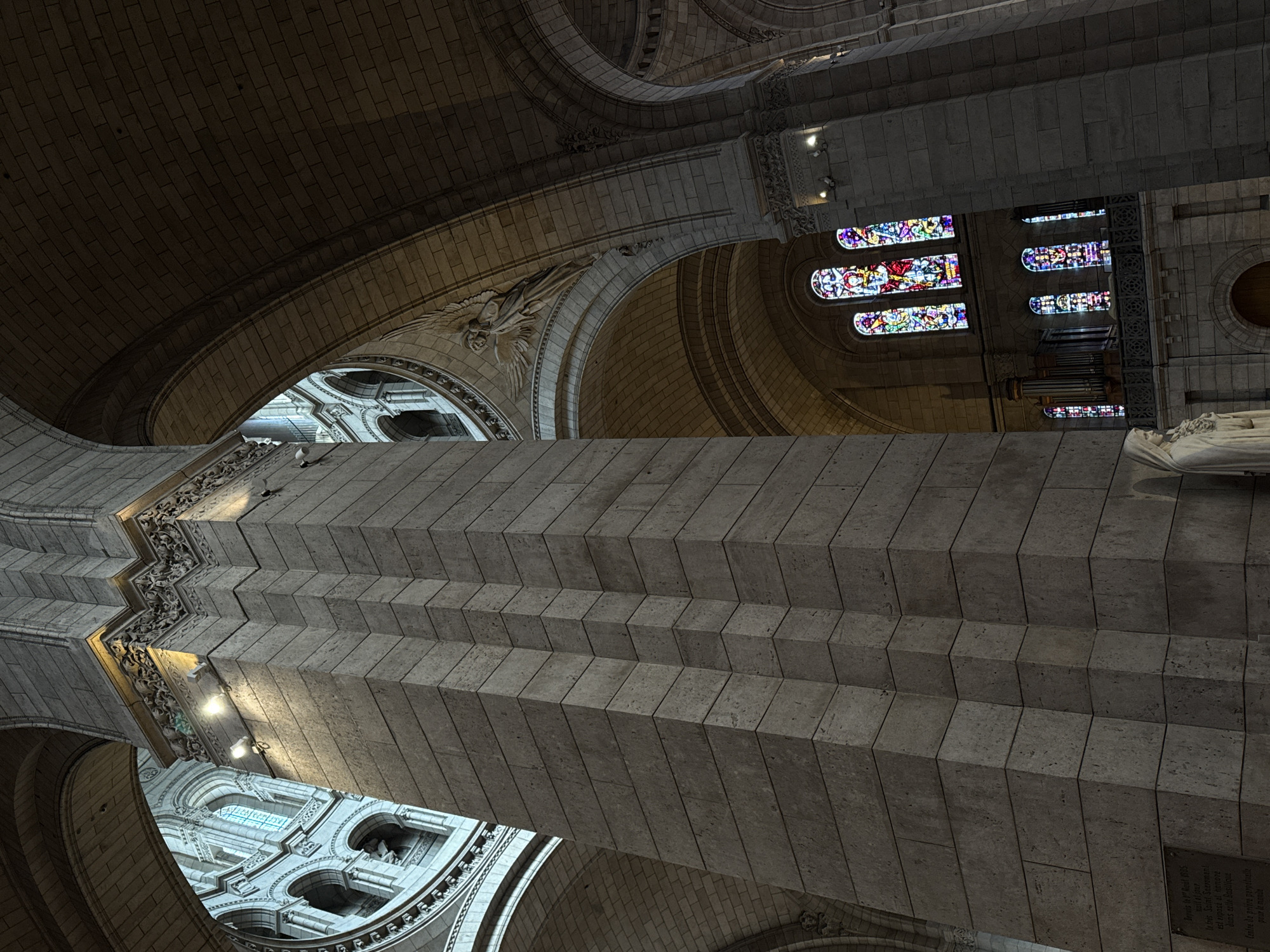 The image shows an upward view of the interior of the Sacré-Cœur Basilica, located in Paris, France. The architectural details are prominent, including stone arches, a multi-tiered structure, and stained-glass windows. A plaque with French text is visible on the right side. The photo captures the grand scale and ornate design of the basilica's interior.