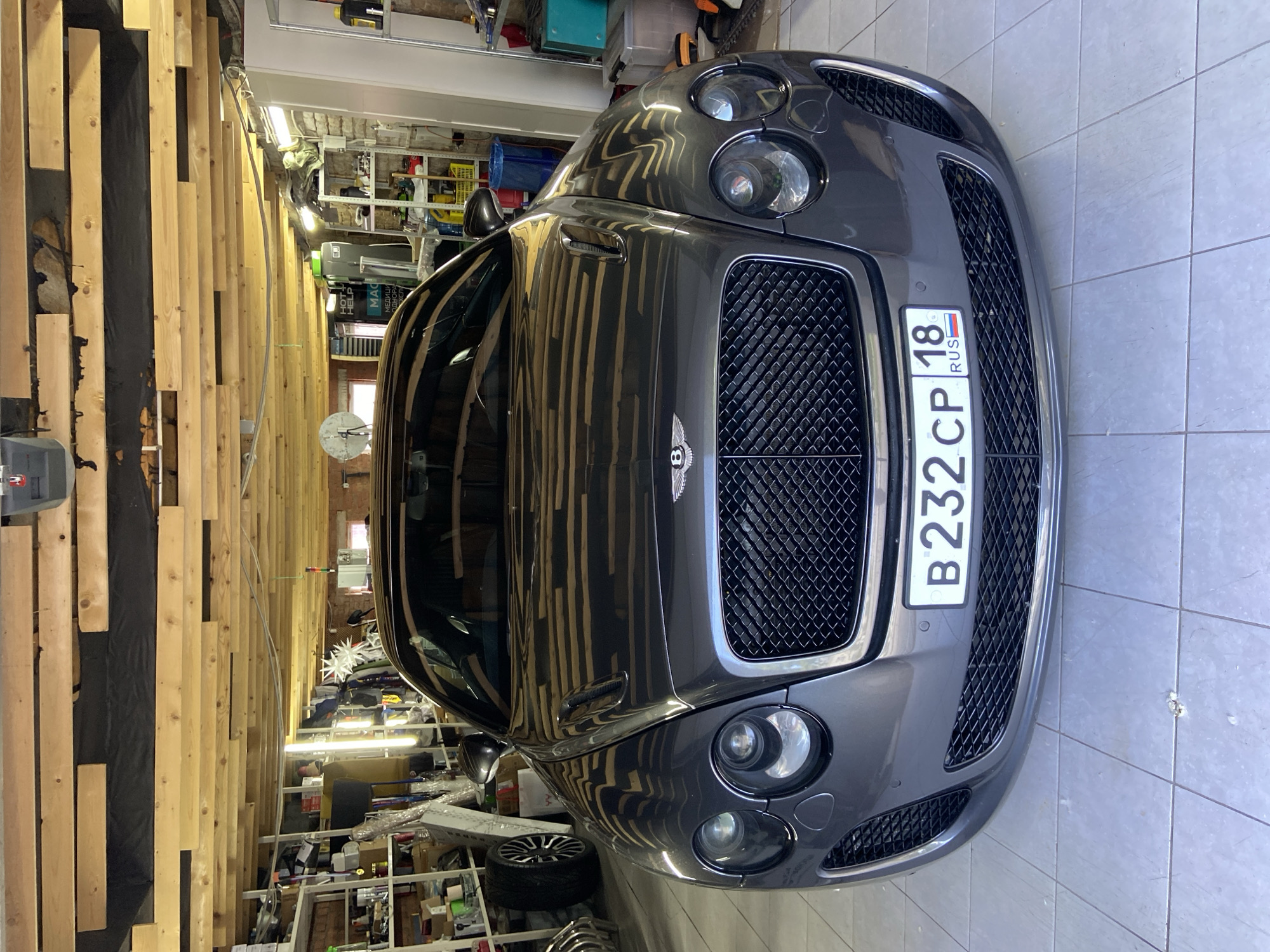 The image shows a grey Bentley Continental GT parked in what appears to be a garage or workshop. The car is positioned head-on, facing the viewer. The garage has a wooden ceiling with exposed beams and lighting fixtures. Shelves filled with various items, including what looks like car parts and cleaning supplies, are visible in the background on the left and right sides. A blue bucket and a white door or panel are also present on the right. The license plate on the car reads "B 232 CP 18 RUS". The floor is tiled. There are no people visible in the image. The image does not provide any information to specifically locate it in the city of Votkinsk, Russia.