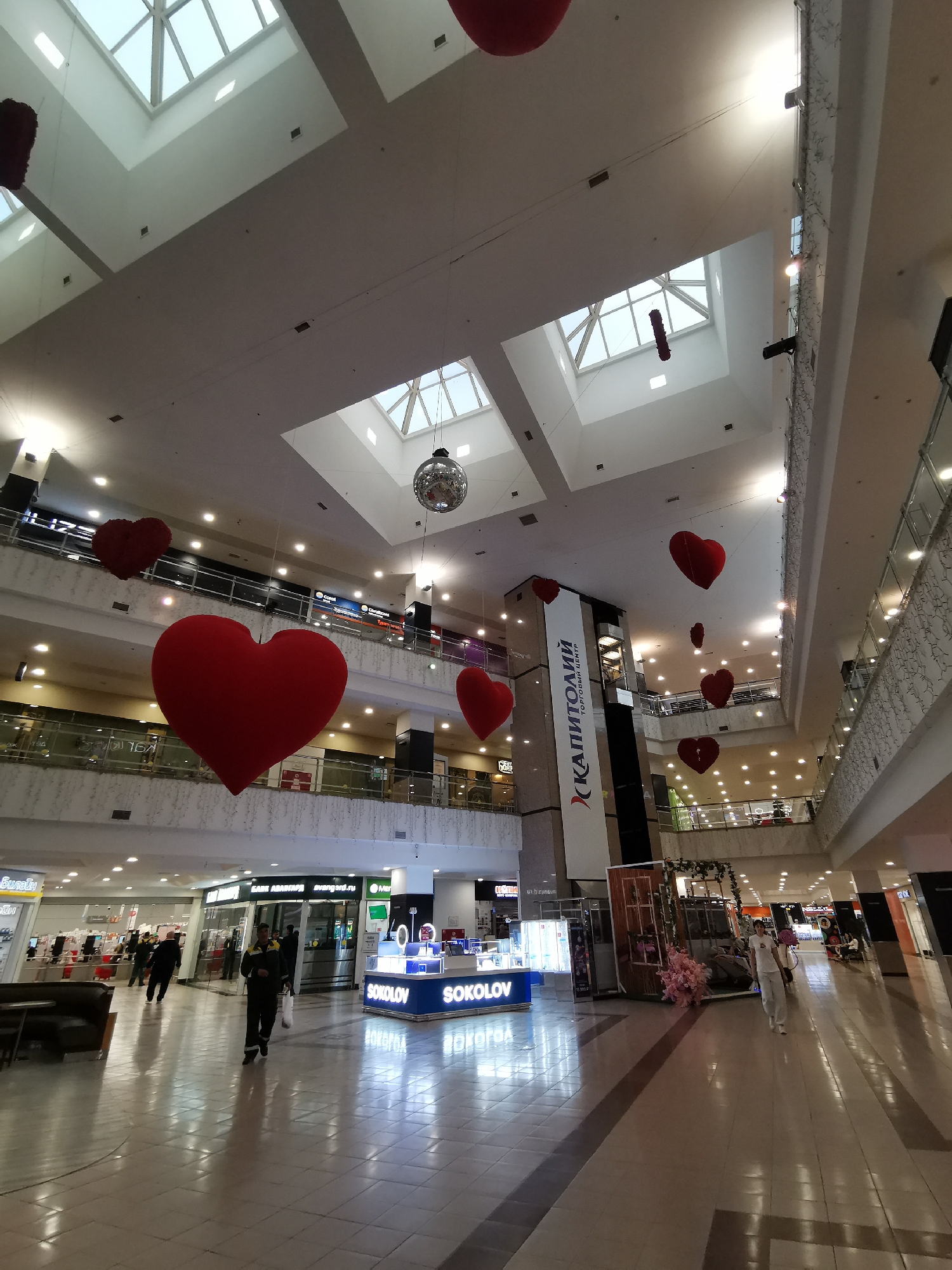 The image shows the interior of a shopping mall in Moscow, Russia, decorated with large, red heart-shaped balloons suspended from the ceiling. The mall has multiple levels, visible through open railings. Several skylights provide natural light. Retail stores line the walkways, with shoppers visible in the foreground and background. One store is identified as "SOKOLOV". A banner for "КАПИТОЛИЙ" is also visible. The floor is tiled and reflects the light from the skylights and overhead lamps. People are walking through the mall.