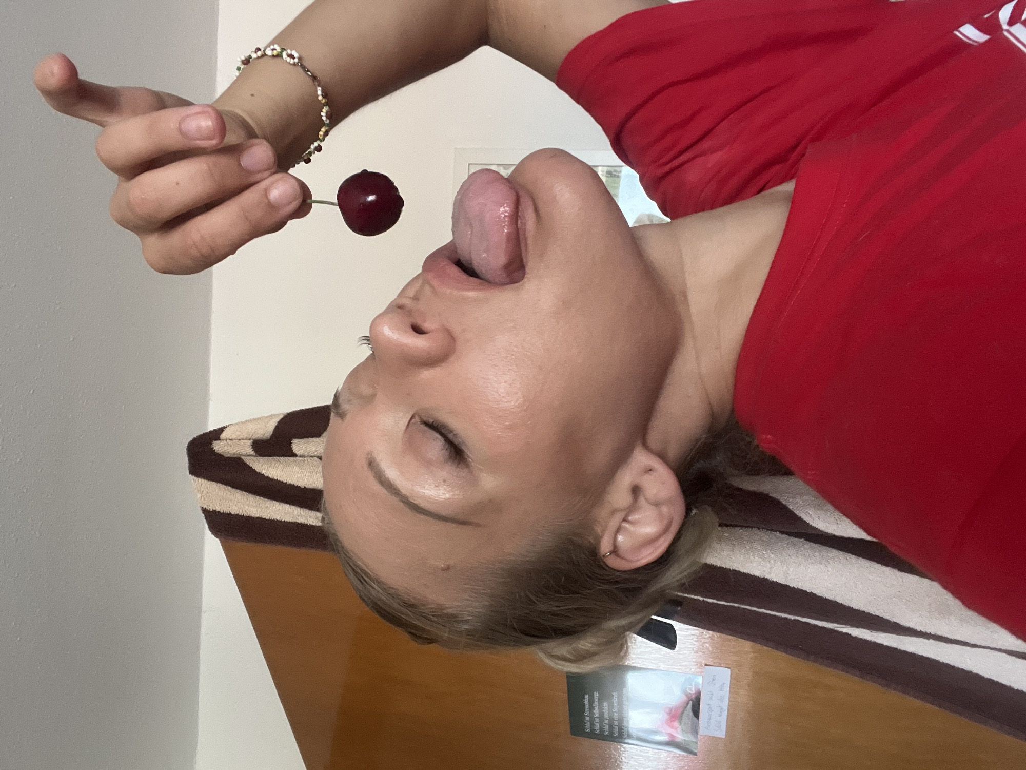 The media file is a photograph featuring a person and a cherry. The person is positioned such that their face is close to the viewer, with their eyes closed and tongue extended to lick a cherry held by their hand. The person is wearing a red shirt. A bracelet adorns their wrist. The background includes a wall and a surface covered with a striped towel. A small booklet with text is also visible. The lighting suggests an indoor setting. The activity captured is the person interacting with the cherry.