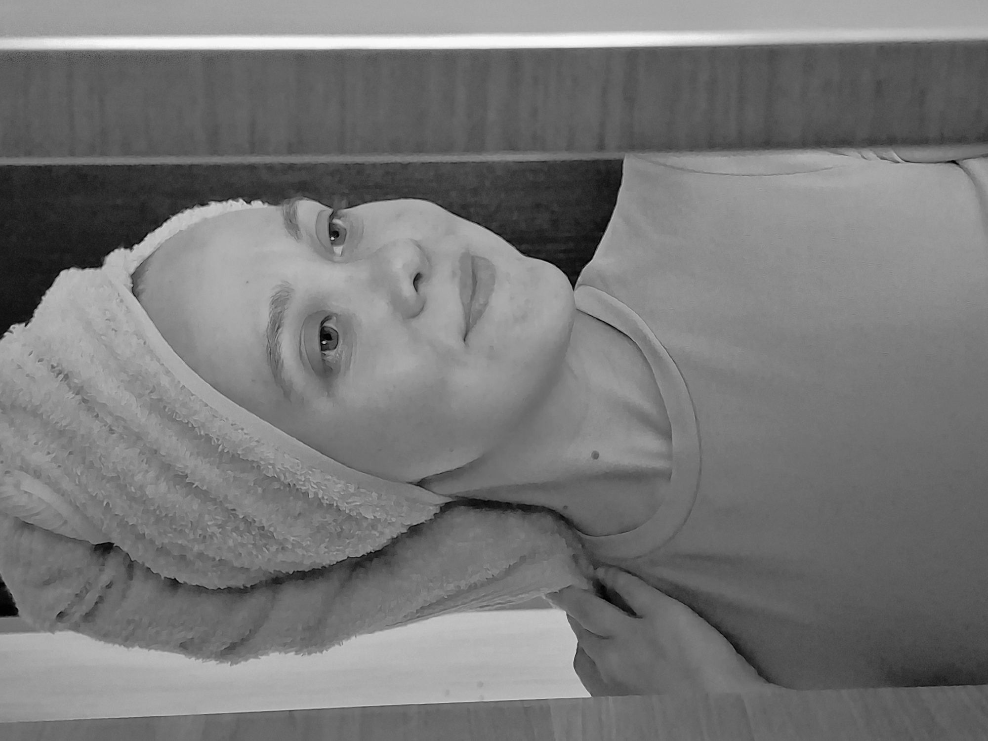 A black and white medium shot shows a person lying down with a white towel wrapped around their head. The person is wearing a light-colored T-shirt. Their face is turned towards the camera, and they appear to be looking slightly upwards. A hand is visible near the bottom right of the frame. The background consists of horizontal lines, possibly a wooden structure. There is no information to indicate that the location is in the city of Chaikovsky, Russia.