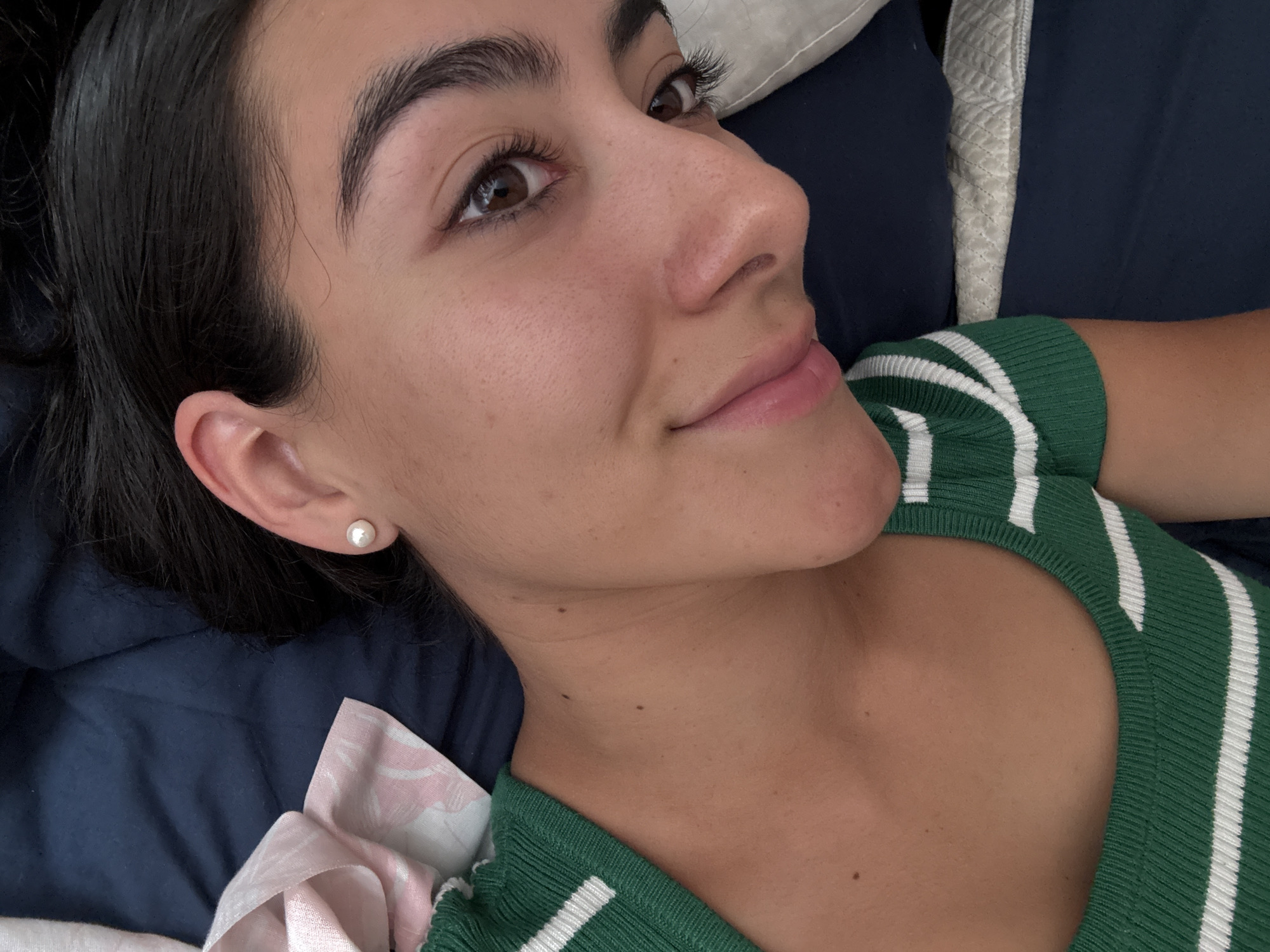 A young woman with dark hair is lying on a bed or surface covered with dark blue fabric. She is wearing a green and white striped top. She is wearing a pearl earring in her left ear. She appears to be indoors, in a room with pillows in the background. The location cannot be determined.