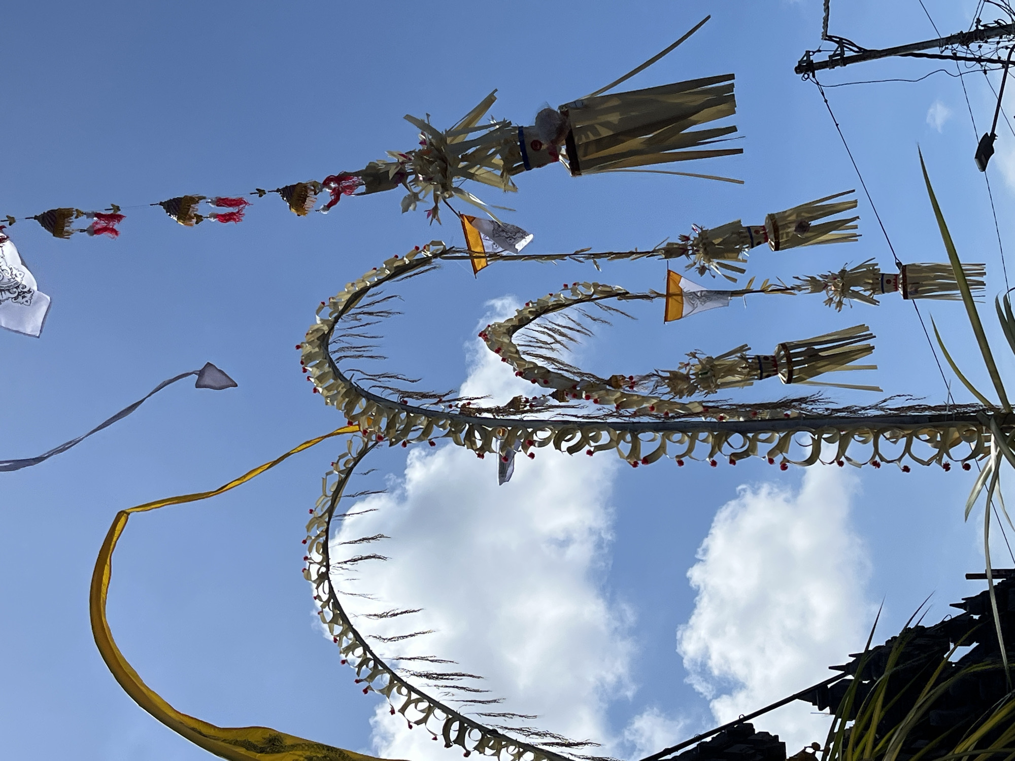 A low-angle shot shows an ornate arrangement of decorations hanging in the air, presumably above a street. The decorations are made of organic materials like bamboo or palm leaves, fashioned into curved shapes and adorned with small flags and dangling ribbons. A long yellow ribbon extends from one of the structures. The background is a bright blue sky with scattered white clouds. Power lines are visible in the upper right corner of the image, suggesting an urban setting. The photograph was taken in Kuta Selatan, Indonesia.