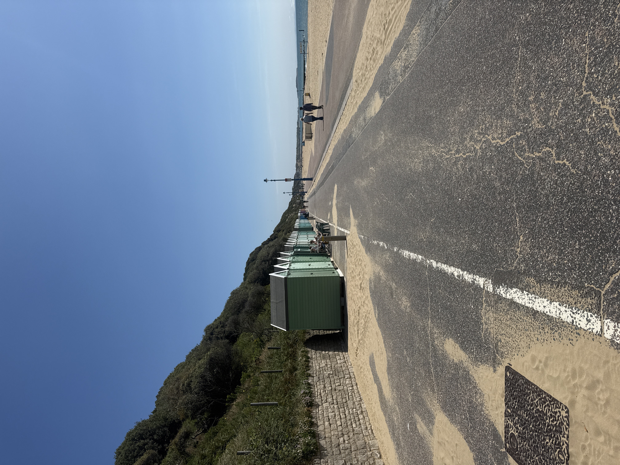 The image captures a sunlit day at a beach promenade, likely in Bournemouth, United Kingdom. The scene is dominated by a clear blue sky. In the foreground, a paved pathway runs diagonally across the frame, bordered by sand on the left and a line of green beach huts on the right. The pathway shows signs of wear, with cracks and sand covering portions of the asphalt. A white line marks the edge of the pathway.
Beyond the pathway, the sandy beach stretches towards the sea, which is visible in the distance under a hazy sky. The beach is mostly empty, with a few people strolling along the promenade in the distance. To the left of the promenade, a lush green hillside covered in dense foliage rises, providing a natural backdrop. A low stone wall runs along the base of the hill, separating it from the promenade.
The beach huts, painted a uniform light green, are neatly arranged along the promenade, their triangular roofs pointing towards the sky. Some appear to be open or have people near them. The overall atmosphere is one of a peaceful, possibly early morning or late afternoon, day at the seaside. The sunlight casts shadows, indicating a bright and clear day. There is no visible text in the image.