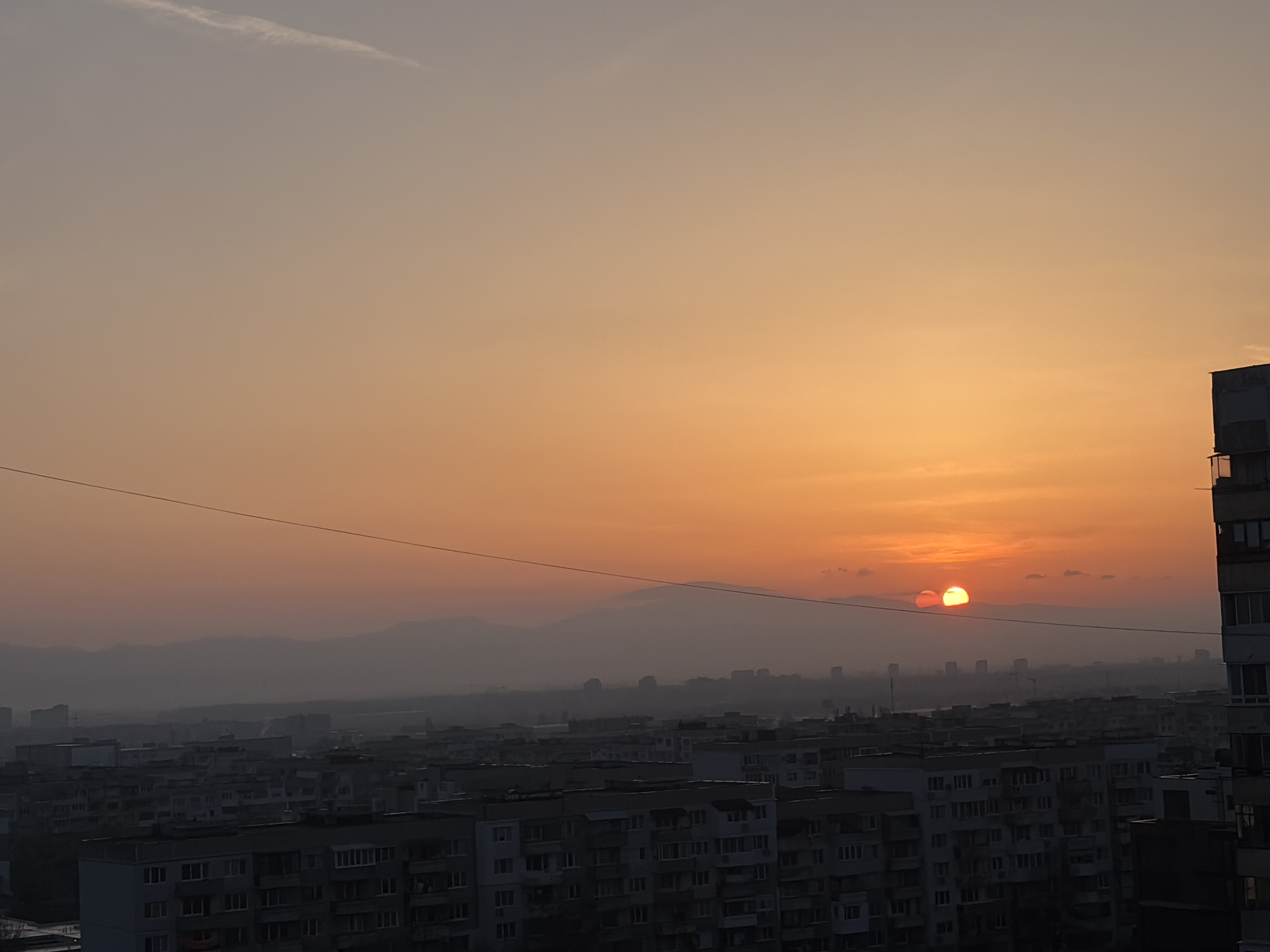 The image presents an urban landscape scene at either sunrise or sunset, viewed from an elevated perspective over Sofia, Bulgaria. The sky occupies the majority of the frame, displaying a gradient of colors from a light greyish-blue at the top to a warm, vivid golden-orange near the horizon. A thin, elongated cloud streak is visible in the upper left portion of the sky.
The sun, a prominent feature, is an intense orange-red disk positioned close to the horizon. It appears distinctly split into two overlapping halves, possibly due to atmospheric refraction, partial obscuration by a thin cloud layer, or lens effects. Small, wispy clouds are clustered around the sun.
Below the horizon, a sprawling cityscape extends across the mid-ground and foreground. The area is densely packed with numerous multi-story apartment buildings, featuring typical concrete block architecture with many windows and balconies, common in Eastern European cities. These structures are largely silhouetted against the bright sky, rendered in dark grey and muted tones. A thin, dark utility cable or wire runs horizontally across the upper section of the urban panorama.
In the distant background, hazy mountain ranges are visible, their contours softly defined and partially obscured by atmospheric haze or mist. The weather conditions appear to be hazy, which contributes to the diffused quality of light and the subdued appearance of distant elements. There are no people or discernible text within the image. The scene conveys a sense of tranquility and vastness, characteristic of dawn or dusk over a bustling metropolitan area.
