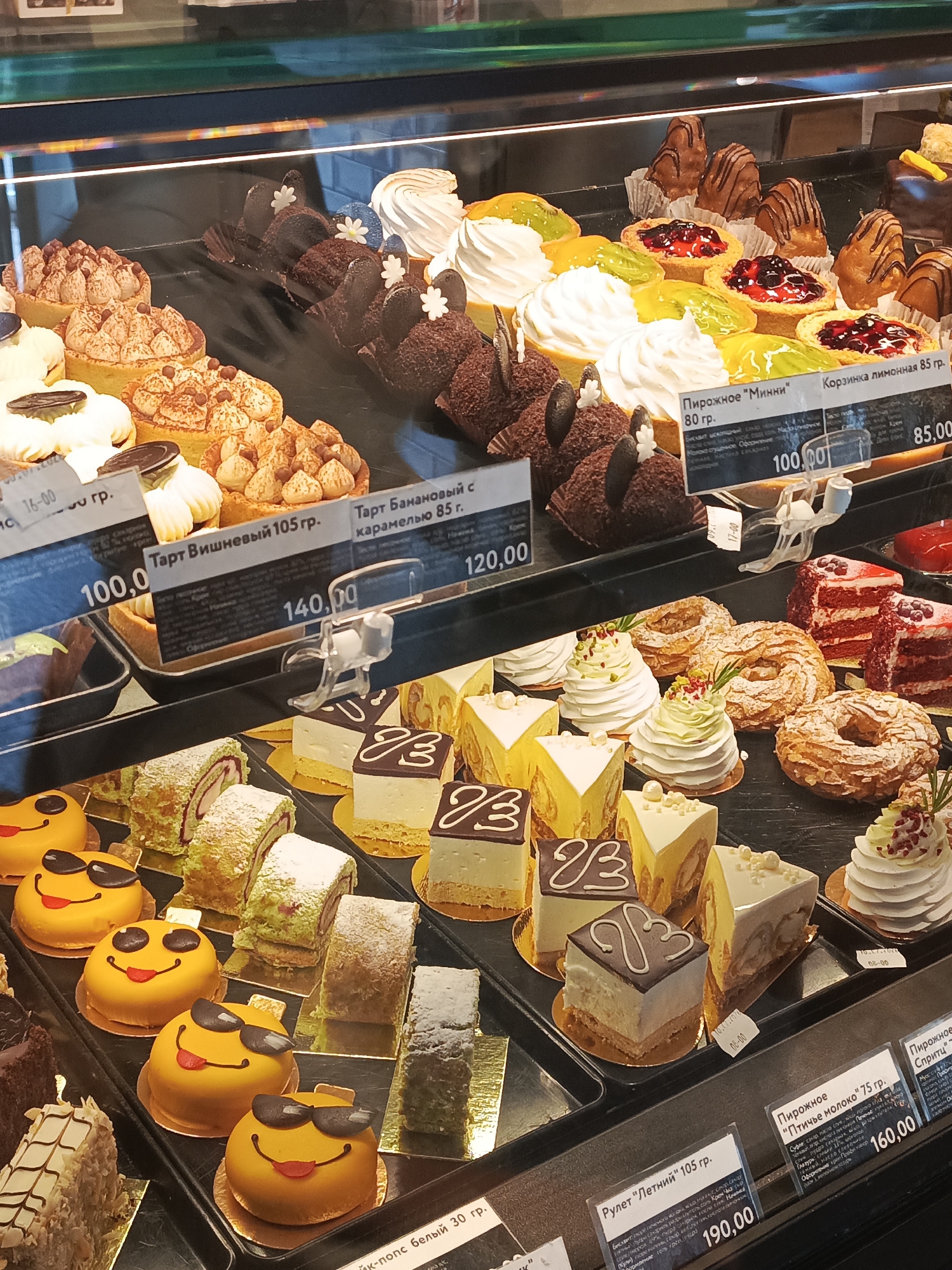This is a close-up, eye-level shot of a bakery display case filled with various pastries and cakes, viewed from the front. The lighting is bright, coming from overhead, creating reflections on the glass. The display is organized into tiers, showcasing a wide assortment of desserts.
In the top tier, there are several small tarts and cakes, some topped with whipped cream and fruit, others with chocolate and nuts. Below this, a row of chocolate-covered cakes with a rough texture is visible. Further down, several yellow, smiley-faced cakes with sunglasses sit in a row, appearing to be part of a children's selection. Behind these, a variety of rolled cakes and rectangular pastries are displayed.
In the middle section, a prominent sign indicates "Пирожное "Минни" 80 гр." (Minnie Mouse Cake 80g) with a price of "100,00". Next to it, another sign reads "Корзинка лимонная 85 гр." (Lemon Basket 85g) priced at "85,00". There are also many other pastries including what appear to be cream puffs, layered cakes with red and white frosting, and ring-shaped pastries with nuts.
In the lower sections, more rectangular and triangular cakes are visible, along with a sign for "Рулет "Летний" 105 гр." (Summer Roll 105g) priced at "190,00". Another sign indicates "Пирожное "Птичье молоко" 75 гр." (Bird's Milk Cake 75g) with a price of "160,00".
The overall impression is of a well-stocked and diverse pastry selection in a clean and brightly lit environment. The text on the price tags and labels is in Russian. The image likely depicts a scene in a bakery located in the city of Kurgan, Russia, as suggested by the Russian text and common bakery offerings found in Russian cities. No people are visible in the image.