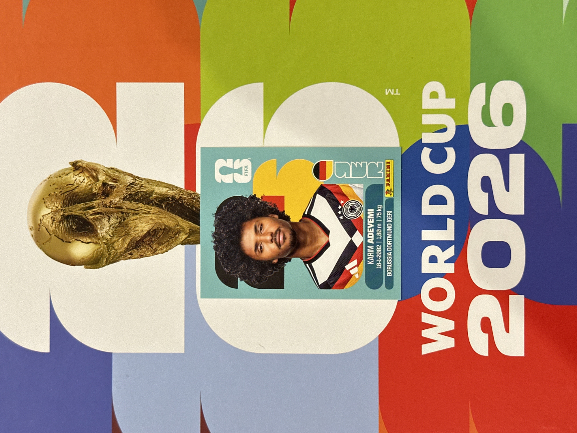 The image displays a vibrant, vertically oriented graphic design centered on the theme of the FIFA World Cup 2026. The background consists of large, overlapping, stylized numerical digits "2026" rendered in a multi-colored geometric pattern, featuring blocks of white, orange, blue, green, red, and light blue.
In the upper central portion of the image, the golden FIFA World Cup trophy is prominently featured, with its upper half visible. Centered below the trophy is a rectangular football (soccer) player's card or sticker. The card features a headshot of a male player, identified as Karim Adeyemi, with dark curly hair, a beard, and a mustache. He is wearing a black and white striped jersey with elements of the German flag colors and the German national team crest on the left chest. Printed details on the card include "KARIM ADEYEMI", "18-1-2002", "1,80 m | 75 kg", and "BORUSSIA DORTMUND (GER)". The card also incorporates a stylized FIFA logo (labeled '23') in the upper right, a vertical strip of the German flag on its right edge, and a Panini logo at the bottom right.
Below the player's card, large white capital letters spell "WORLD CUP", followed by the prominent white digits "2026", which align with the stylized numbers in the background. A "TM" (trademark) symbol is partially visible in the lower right quadrant. The image contains no visual elements or contextual information pertaining to a specific geographical location such as the city of Prisovo, Bulgaria.