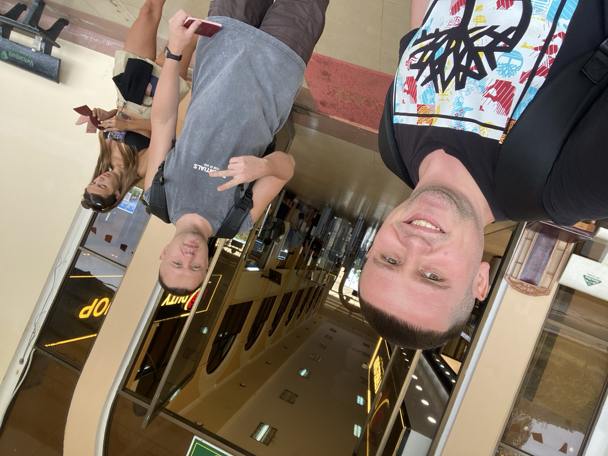 Three individuals, two males and one female, pose for a selfie outside a duty-free shop in the city of Bo Y, Vietnam. The male in the foreground, wearing a black t-shirt with a graphic print and a backpack, smiles at the camera. To his right, another male in a gray sleeveless t-shirt and brown shorts holds up two fingers in a peace sign and is also holding a passport. Behind them, a female wearing a black bikini top and a tan skirt looks down at a passport in her hands. The background features the entrance to a duty-free shop with reflective windows and a yellow-toned interior. A bench with the "Vietcombank" logo is visible to the right.