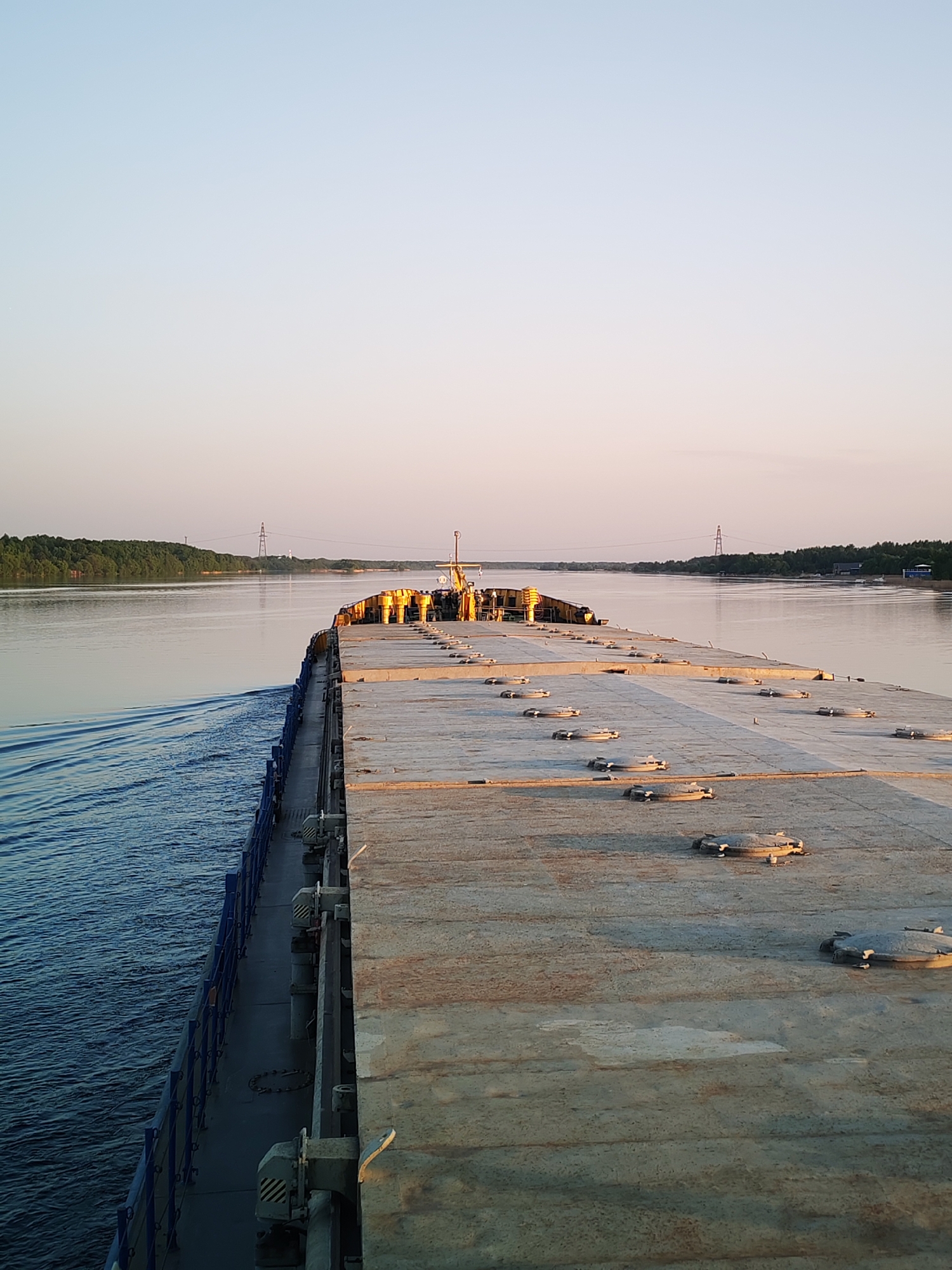 The image shows a view from the deck of a barge on a wide river. The barge is carrying a deck of square or rectangular concrete-like panels, each with circular hatches. The deck appears aged and worn. The river stretches out towards the horizon, flanked by tree-lined shores on either side. In the distance, electrical transmission towers are visible. The sky is a pale blue, suggesting either early morning or late evening light. The river water is relatively calm, with some visible wake trailing behind the barge on the left side. This scene is in Bely Gorodok, Russia.