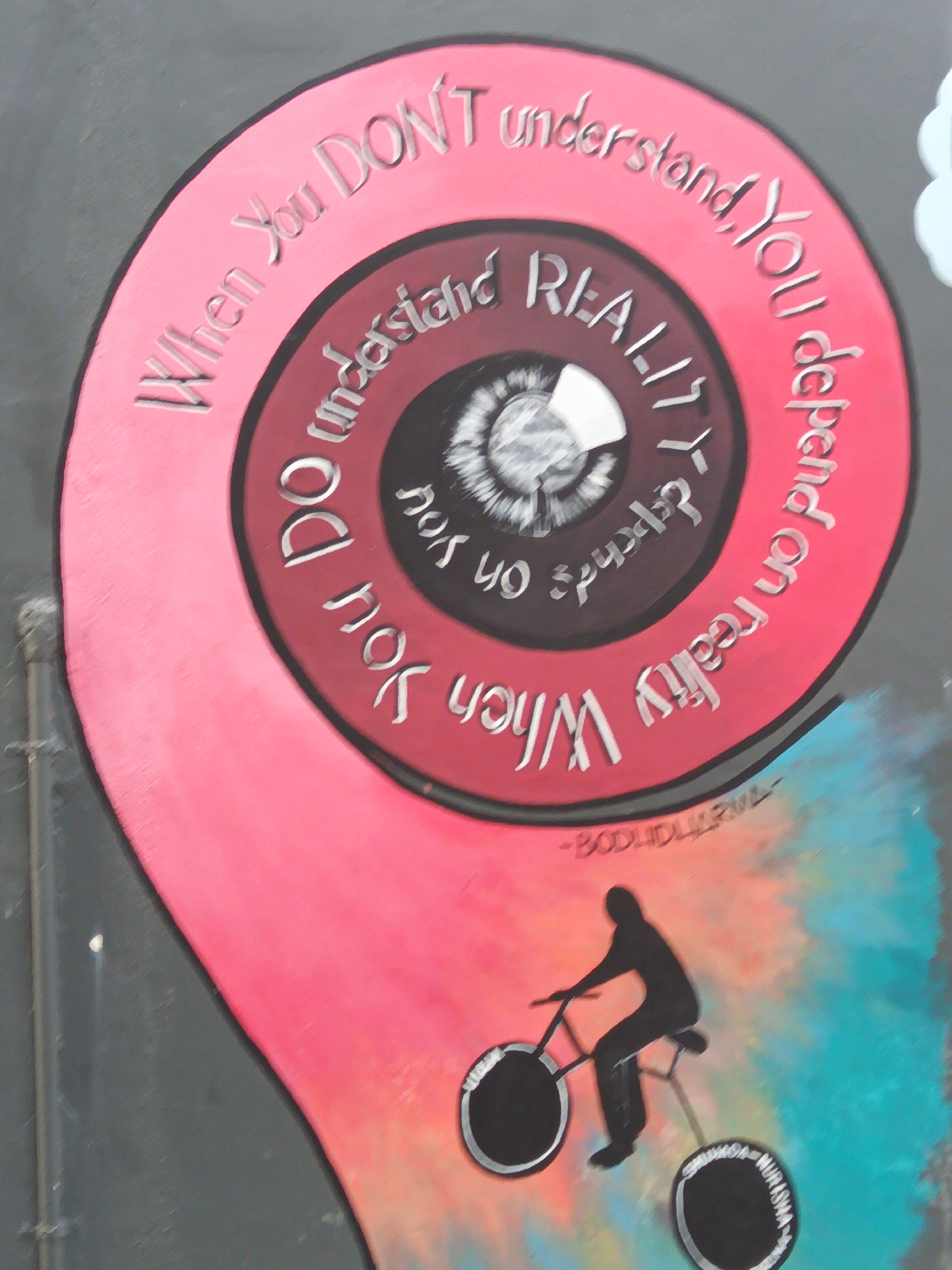 A mural painted on a wall in Kuala Lumpur, Malaysia, depicts a spiral with text. The text reads, "When You DON'T understand, YOU depend on reality. When YOU do understand REALITY, YOU do not depend on YOU." A silhouette of a person riding a bicycle is shown below the spiral. The bicycle wheels also have text inscribed on them, partially obscured. The colors used in the mural are pink, red, black, white, and teal. The background wall appears to be grey. The mural has a textured, carved appearance.