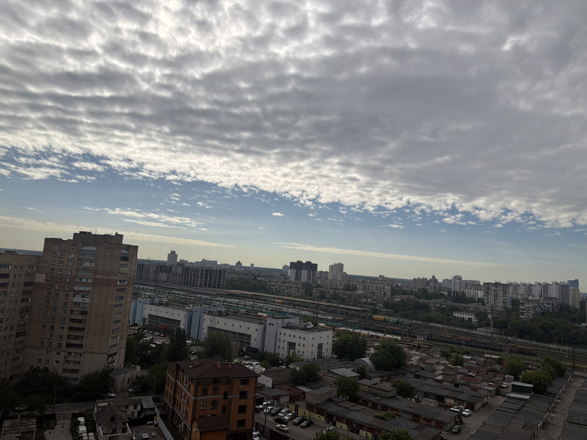 The image depicts an aerial view of an urban landscape in Kyiv, Ukraine, likely taken from a high vantage point. The foreground features a dense collection of low-rise buildings, some appearing to be garages or workshops with corrugated metal roofs. Further back, a large, multi-story residential building dominates the left side of the frame, characterized by its pale beige facade and numerous balconies. Beyond this, a sprawling railway network is visible, with multiple tracks and numerous train cars, suggesting proximity to a major transportation hub. In the mid-ground and background, a cityscape unfolds with a variety of architectural styles, including numerous high-rise apartment buildings and other structures. The sky is partly cloudy, with textured grey clouds spread across a blue sky. The overall impression is that of a densely populated urban environment with a significant industrial or transportation presence.