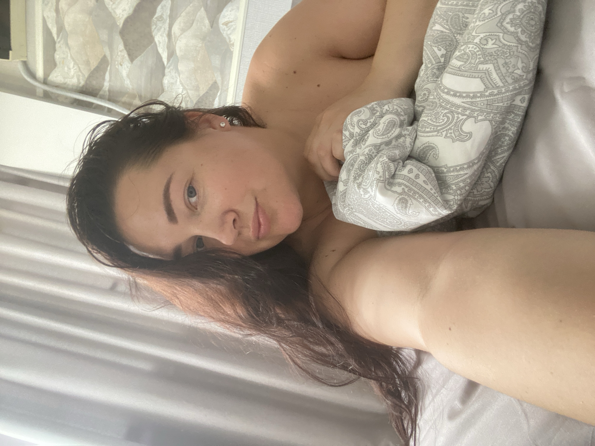 The image depicts a young woman lying in bed, with her head propped up on pillows and her body partially covered by a duvet. She has long, dark brown hair that cascades over her shoulder and a fair complexion. Her eyes are blue, and she is wearing small, sparkly earrings. The background shows a light-colored curtain on the left and a patterned wallpaper with a light-colored design on the right, suggesting an indoor setting, likely a bedroom. The lighting appears to be natural, possibly from a window. The overall mood is relaxed and intimate. The location is not specified in the image or the provided crops, and therefore, cannot be identified as Sochi, Russia.