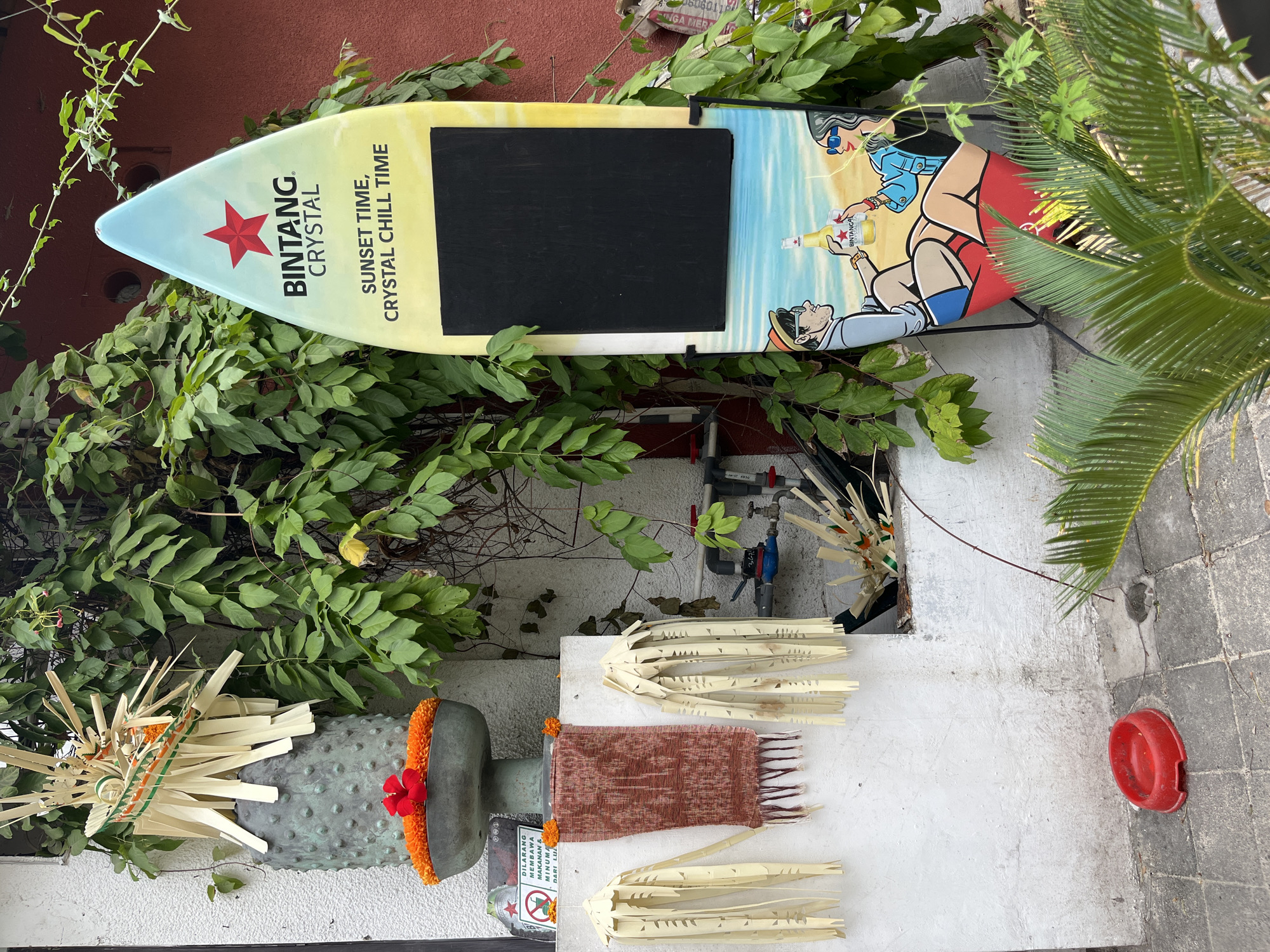 An outdoor scene in Kuta, Indonesia, features a vertical surfboard-shaped sign for "BINTANG CRYSTAL" positioned on the right. The sign displays text that reads "SUNSET TIME, CRYSTAL CHILL TIME" and has a black rectangular section for writing. Below this, a cartoon depicts a man and a woman on a beach, toasting with Bintang beer. To the left of the sign, a white wall is adorned with Balinese decorations, including what appear to be offerings of flowers and woven palm leaves. Hanging from the wall are two sets of elaborate, fringe-like palm leaf decorations. A rustic, woven textile hangs between them. In the foreground, a red pet bowl rests on a paved surface. Lush green foliage surrounds the scene, with a portion of a red building visible in the background.