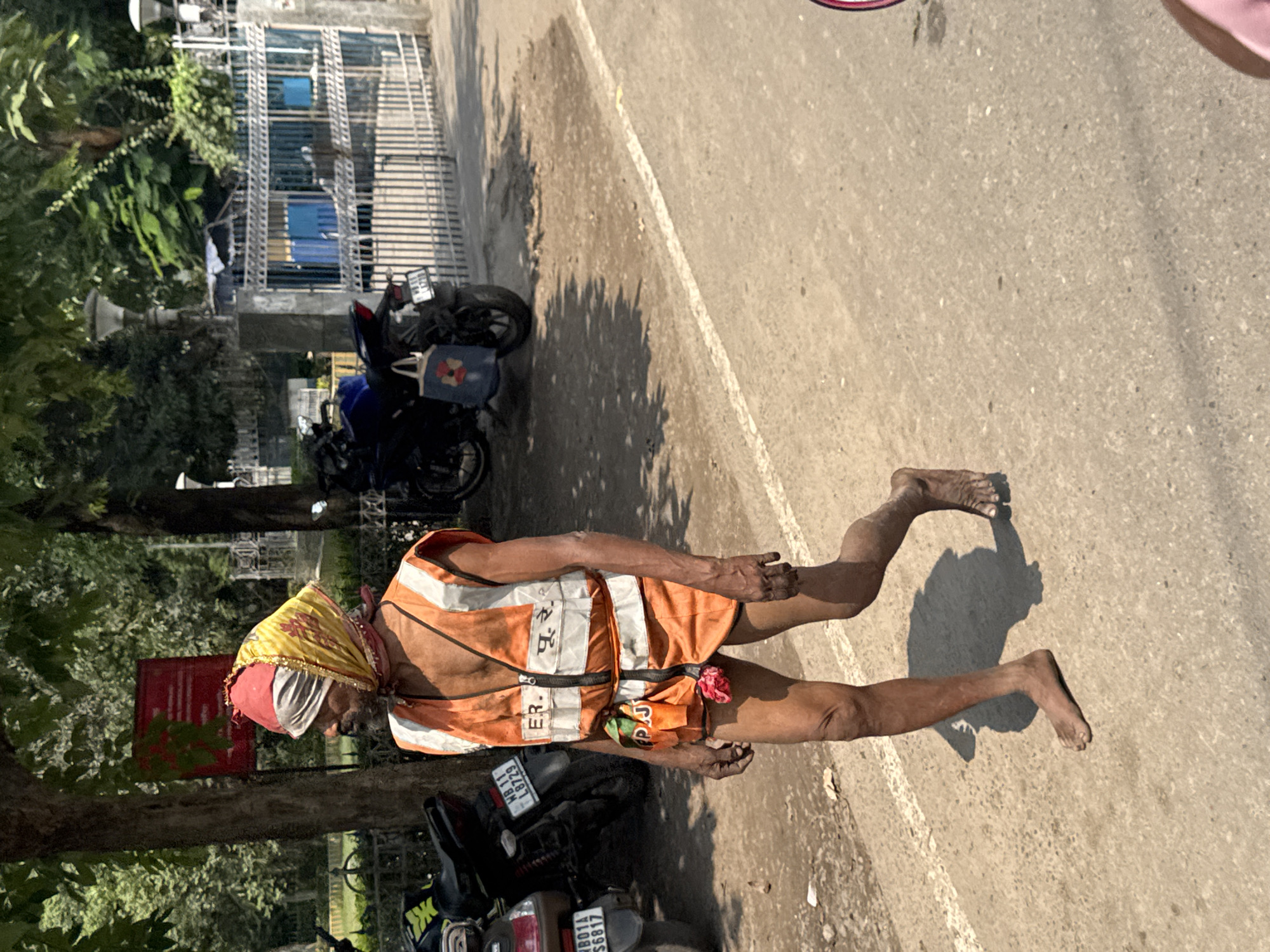 A person wearing a saffron head wrap, an orange and white vest, and nothing else is walking barefoot on a gray asphalt road. The person is tan-skinned and appears to be elderly. Their left arm and hand are partially obscured by the vest, while their right arm hangs at their side. The road is in a state of slight disrepair. To the left of the person is a tree trunk. Behind the tree are two parked motorbikes and a metal fence. Further back, a building can be partially seen. The location is potentially Kolkata, India.