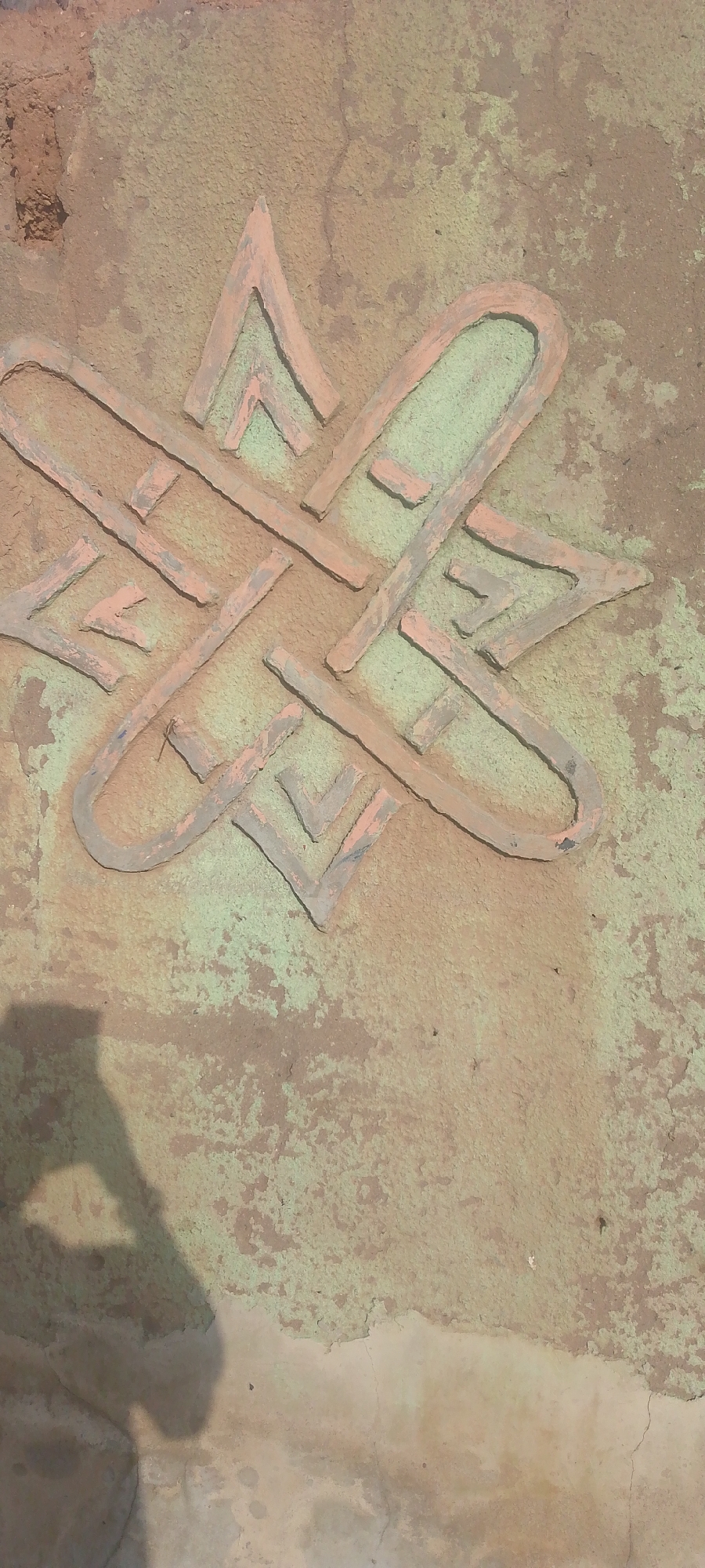 This is a close-up, high-angle shot of a wall with an embossed design, likely in Jalingo, Nigeria. The design is geometric, appearing as an interwoven star or knot pattern. The embossed lines are a light peach color, contrasting with the textured wall, which is a mottled mix of earthy brown and a greenish-gray, suggesting age and weathering. The surface of the wall shows signs of cracking and chipped plaster, further indicating its age or outdoor exposure.
In the lower left corner of the image, a shadow of a person is visible, cast onto the wall. The shadow suggests the presence of a person, possibly the photographer, standing with their left side towards the wall. The lighting and the sharp edges of the shadow imply that the photograph was taken during the daytime, likely with bright sunlight. No specific weather conditions are discernible, though the texture of the wall might suggest it has been exposed to the elements. No text is visible in the image. The overall impression is one of an aged, textured surface with a decorative element, set against the backdrop of a possibly rural or older urban environment in Nigeria.