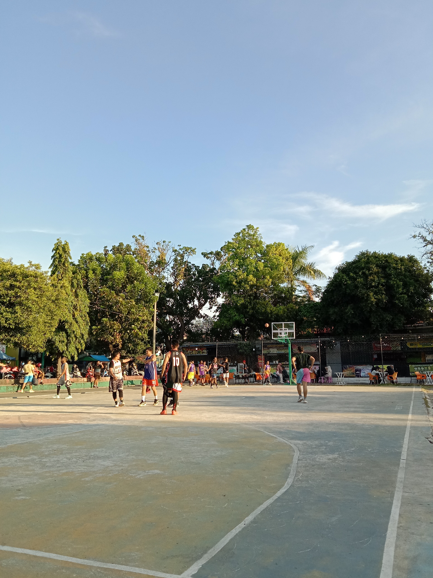 The image depicts an outdoor basketball court in Lhokseumawe, Indonesia. Several people are on the court, some wearing basketball jerseys and shorts, engaged in a game or practice. One person is positioned near the basketball hoop, appearing to have just released a ball. Additional individuals are present around the perimeter of the court, either watching the game or engaged in other activities. Trees and building structures are visible in the background. The sky is clear with some clouds.