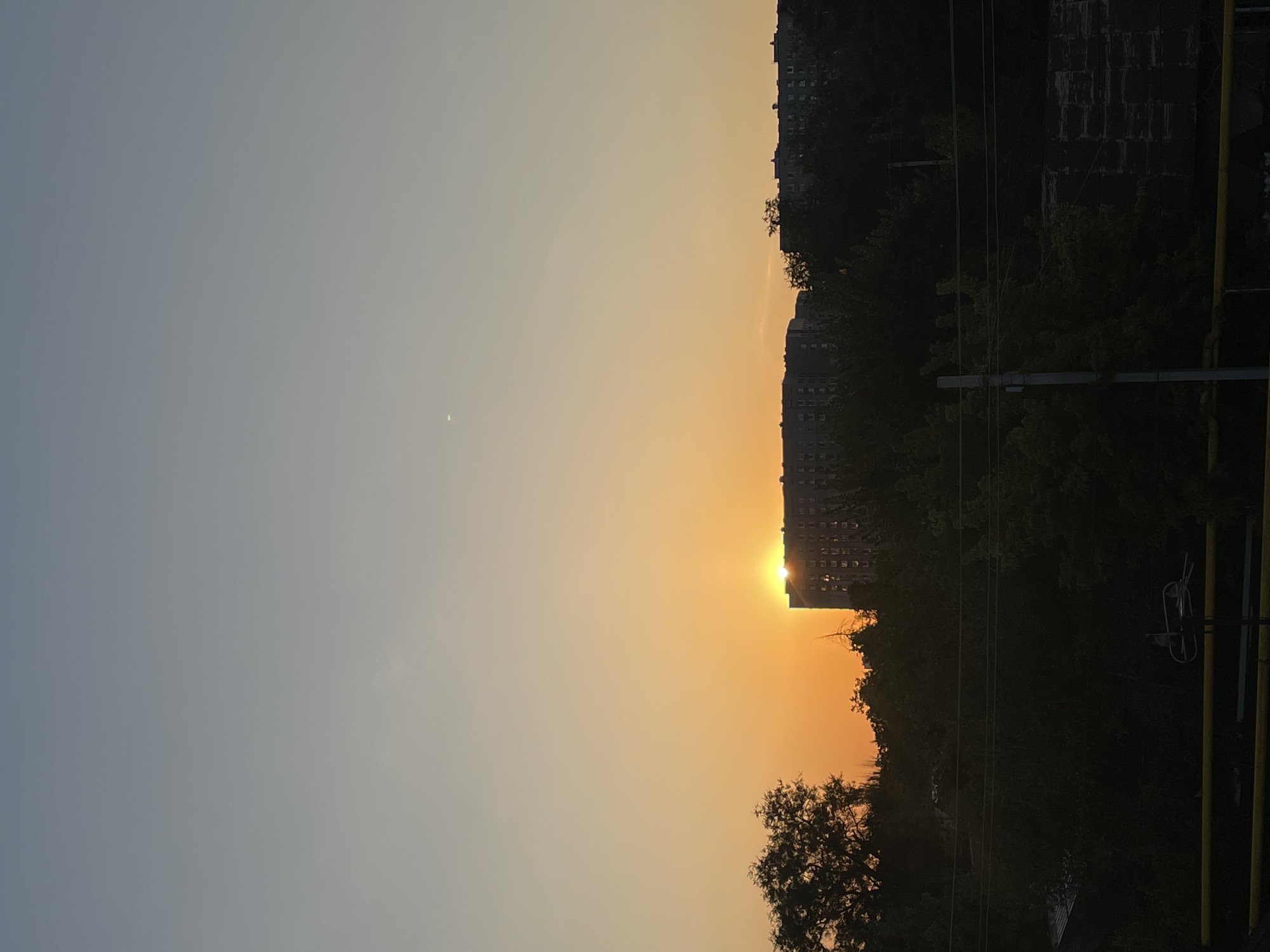 The image depicts a sunset in Almaty, Kazakhstan. The sun is partially obscured by a large, dark apartment building, casting an orange glow across the sky. Silhouetted trees and foliage form a dark band across the middle of the frame. In the foreground, a yellow railing and a dark, indistinct structure are visible. The sky transitions from a pale orange near the horizon to a muted blue-gray at the top of the frame. A tiny white dot, possibly the moon or a distant object, is visible in the upper sky. The scene captures a typical urban sunset in Almaty.