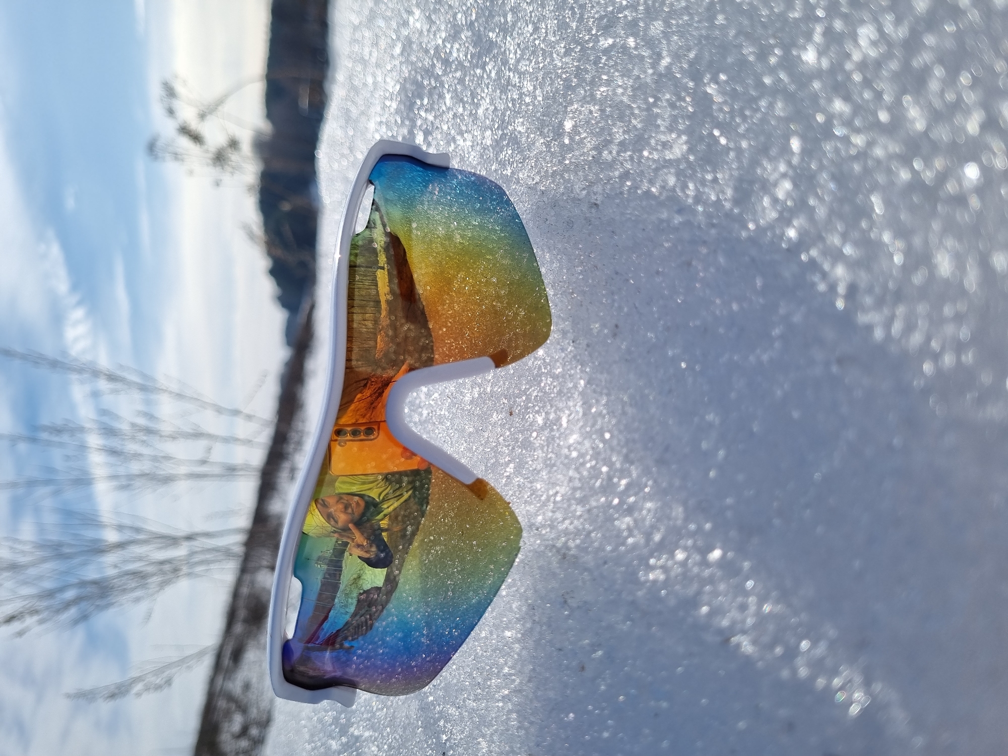 A pair of white-framed sports sunglasses with rainbow-mirrored lenses rests on a granular, crystalline snow surface, which glitters under bright sunlight. Reflected prominently in the left lens is a person, likely female, wearing a yellow beanie and a chartreuse jacket, smiling and making a peace sign with her right hand. Immediately to her right within the reflection, the rear camera array of a mobile phone is visible, indicating it is being used to capture the image. The reflection also shows a snowy ground with what appears to be a fence or structure in the background. Beyond the sunglasses, the environment is a snow-covered landscape under a clear blue sky with some scattered white clouds. Sparse, bare vegetation is visible in the middle ground, indicating an outdoor winter scene in the city of Chaikovsky, Russia.