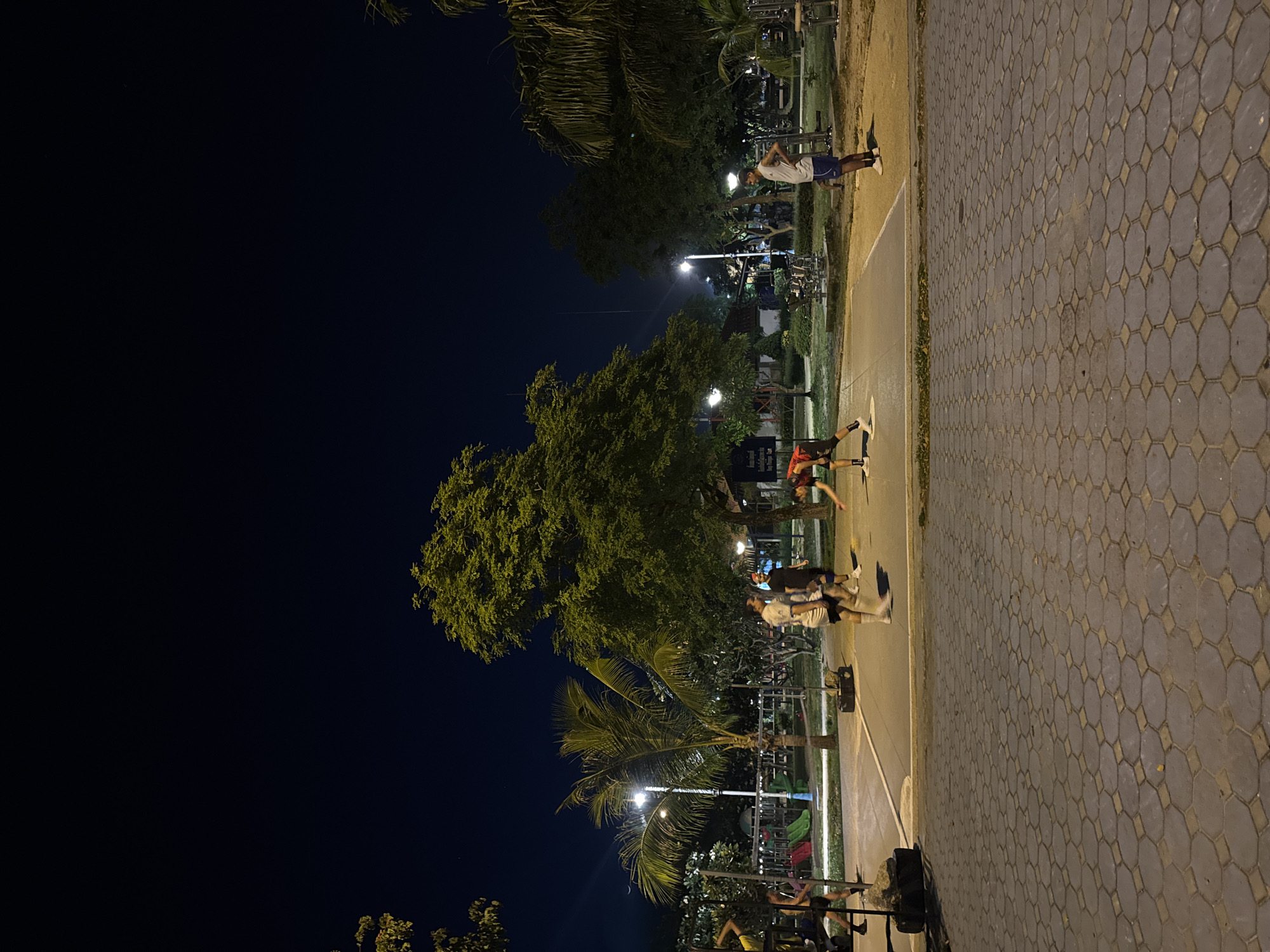 An outdoor basketball court at night in Koh Phangan, Thailand features five individuals. One person in a red and black shirt is bent over on the court, possibly retrieving a ball. Three other individuals, two in white shirts and shorts and one in a black shirt and shorts, stand on the court. A fifth person in a white tank top and blue shorts stands on the edge of the court. The court is adjacent to a playground with slides, and palm trees and other foliage are visible in the background. The ground is paved with hexagonal and diamond-shaped interlocking stones. The scene is illuminated by artificial lights.