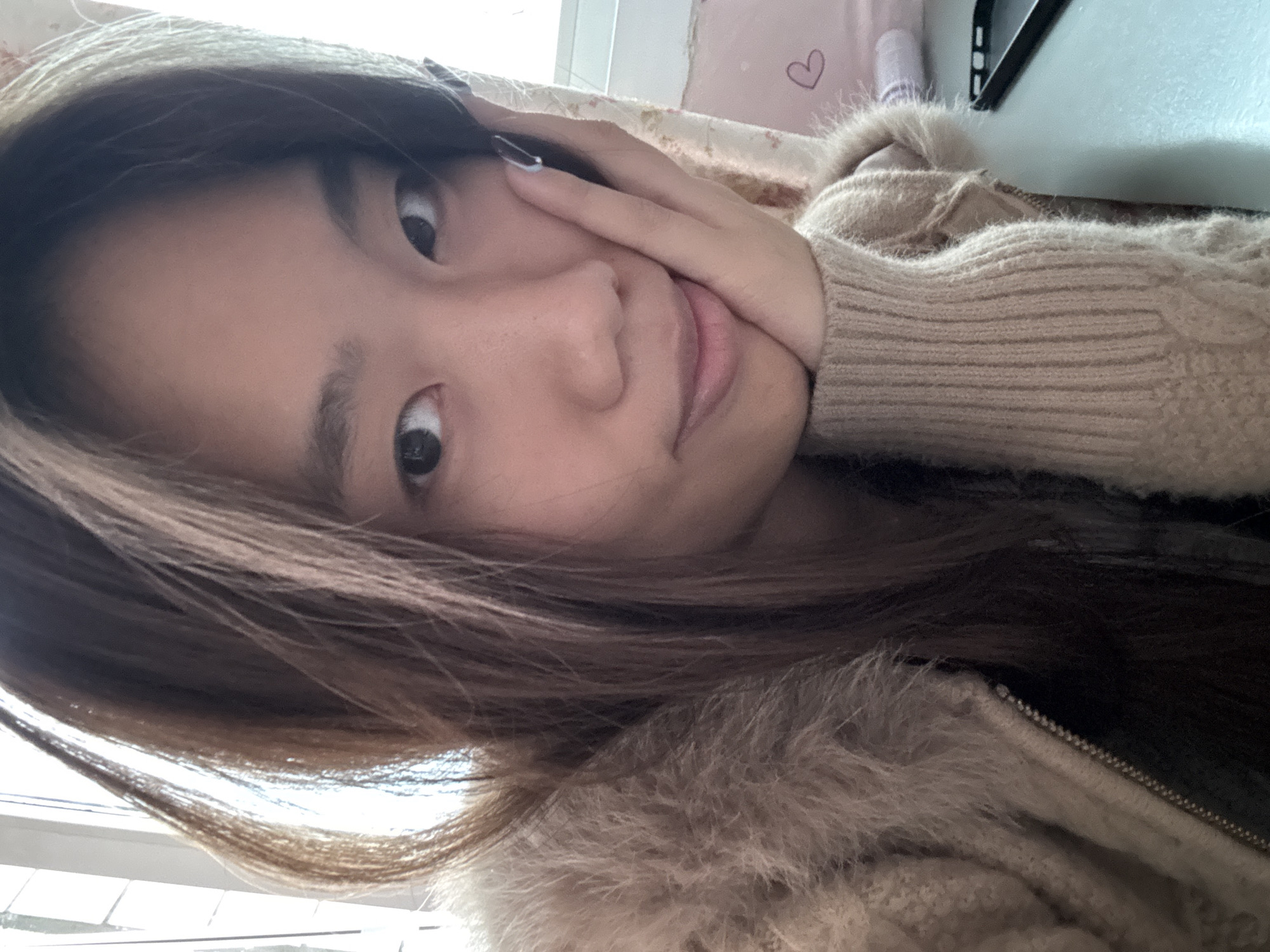 A young Asian woman is pictured indoors, likely in a residential setting. She is wearing a light brown, knitted sweater with a fur-like collar. Her hair is dark brown and partially obscures her face. She is looking directly at the camera, and her hand is touching her face. The background includes a patterned pillow and a window. There is no discernible information to determine the location to be Shapingba District, China.