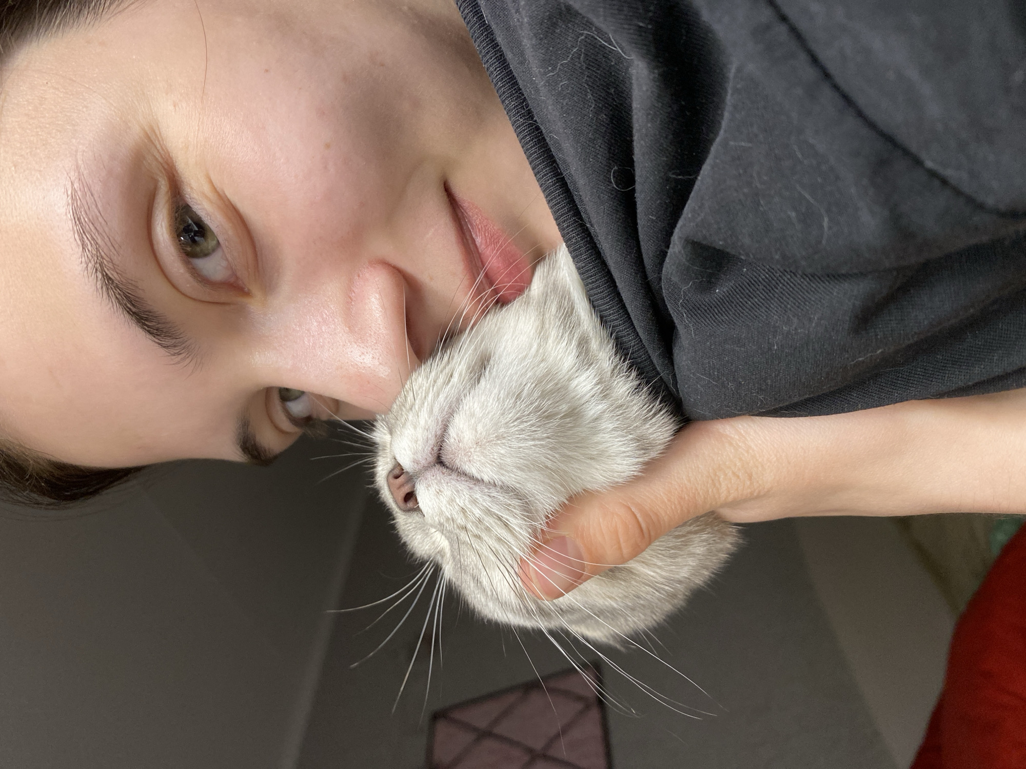 A young person with fair skin and light hair holds a gray cat close to their face. The person's face is turned slightly towards the camera, and their left eye is visible. They are wearing a dark-colored shirt. The cat is being held in their right hand, its nose and whiskers are visible. The background is a dimly lit room. There is no evidence of the location being in Votkinsk, Russia.