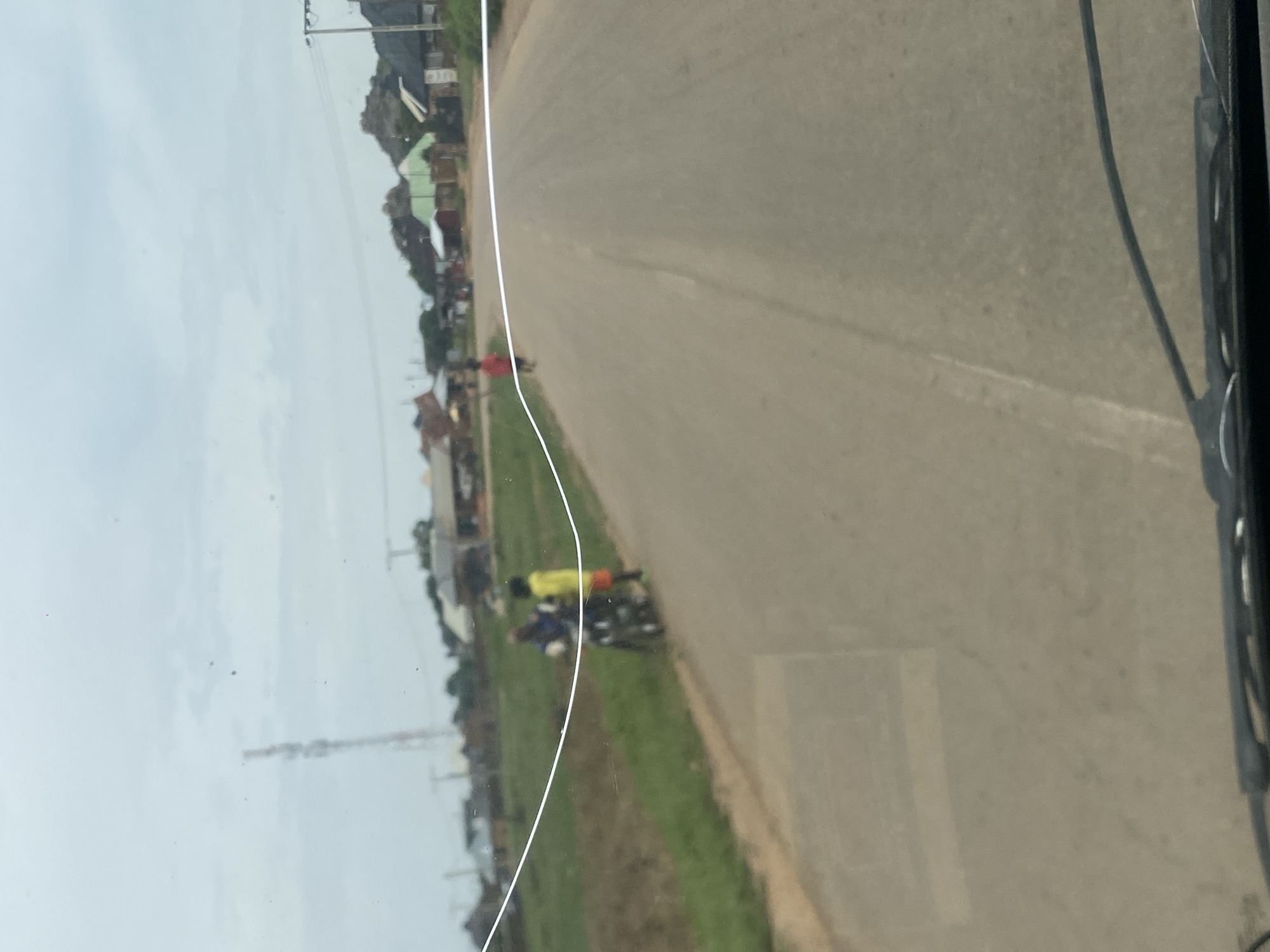 The image depicts a road scene in DU, Nigeria, characterized by a paved road that stretches into the distance under an overcast sky. The road is bordered by a grassy verge on the left side, with signs of agricultural activity in the background. Several structures, appearing to be residential buildings and possibly small businesses, are visible along the sides of the road, with some featuring corrugated iron roofs. A prominent communication tower stands tall on the horizon to the left, a common feature in Nigerian landscapes. In the middle ground, two individuals are present near a motorcycle on the grassy verge. One individual, wearing a blue shirt, appears to be seated on the motorcycle, while another, in a yellow shirt, stands beside them. Further down the road, a person in a red shirt is walking away from the camera. The presence of the motorcycle, the individuals, and the surrounding buildings suggests a typical rural or semi-urban setting in Nigeria. The overall atmosphere is somewhat subdued due to the cloudy weather.