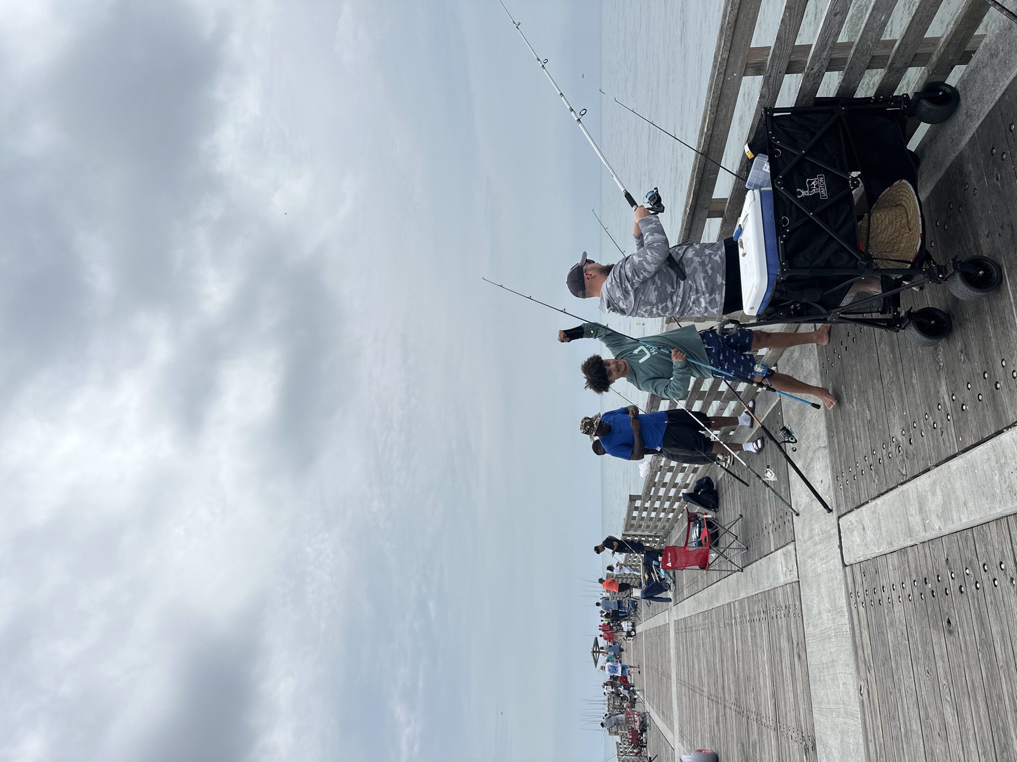 This photo depicts people fishing from a pier on a cloudy day in Jacksonville Beach, United States of America. A man in a camouflage shirt is holding a fishing rod in the foreground. To his left, a younger person with curly hair and wearing a light green shirt and blue patterned shorts is also holding a fishing rod. Another person wearing a blue shirt and black shorts is behind them to the left. A black wagon sits to the right with a cooler, a straw hat, and tackle boxes inside. Fishing poles are propped against the wagon. The pier is made of wood and stretches into the ocean. Other people are fishing farther down the pier. In the background, the sky is overcast.