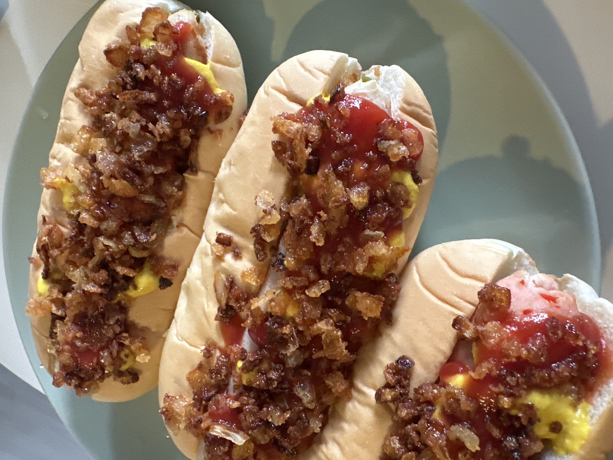 Three hot dogs are displayed on a light green plate. Each hot dog is in a bun and topped with yellow mustard, red ketchup, and crispy fried onions. The hot dogs are arranged diagonally across the plate, with the closest one partially cropped at the bottom. The lighting suggests an indoor setting, possibly a kitchen or dining area. There is no indication of the location being in Kathu, Thailand.