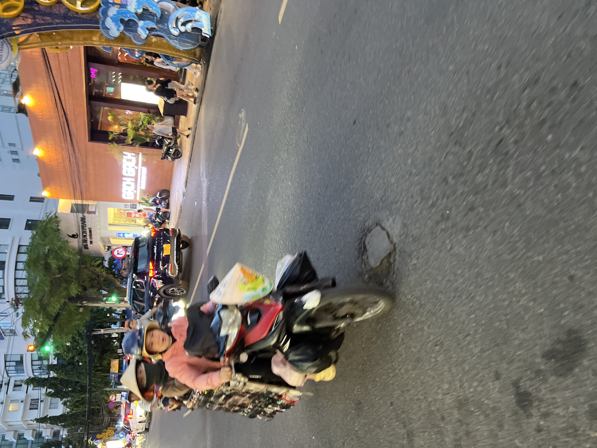 This image captures a street scene in Nha Trang, Vietnam, at what appears to be dusk or early evening. In the foreground, a motorcycle with three occupants is in motion on a paved road. The driver, wearing a helmet and a pink hoodie, is at the front. Behind them, a person wearing a conical hat and a dark top is seated, with a third individual, partially obscured, behind them. The motorcycle is laden with items, including bags on either side and a white plastic bag with a colorful design hanging from the handlebars.
To the left of the motorcycle, a dark-colored SUV is also on the road. Further back, other vehicles, including cars and motorcycles, are visible in traffic. The background features buildings, including a multi-story white building with balconies and a lower, orange-colored building with illuminated signage that reads "BACH GACH" in neon letters. Plants are visible in front of the orange building, and several people are walking on the sidewalk. A decorative structure with blue wave-like elements and yellow accents is positioned on the right side of the frame. Traffic lights are visible on poles, indicating an intersection. The road surface shows some wear and tear, including a patch and some dark stains. The overall impression is a busy urban street during a period of reduced natural light.