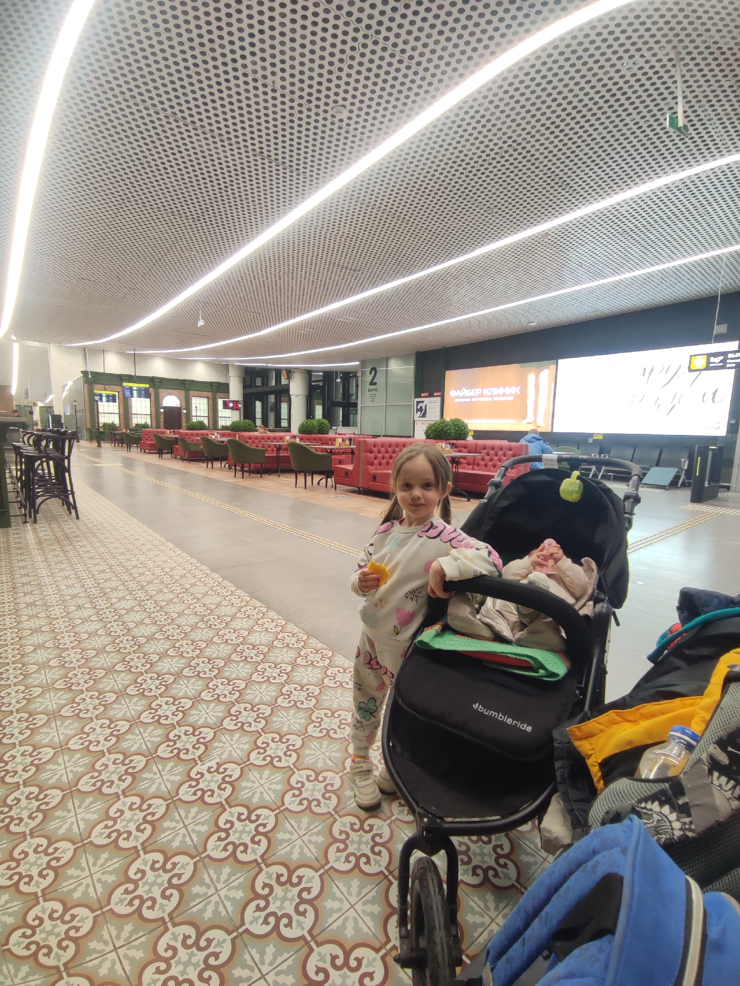 The scene depicts a well-lit, spacious indoor public area, identified as a facility within Izhevsk, Russia. A young female child, approximately 4-6 years old, stands in the foreground, slightly left of center, facing the camera with a slight smile. She has light brown hair styled in two pigtails and wears a white, patterned long-sleeved top and matching pants, along with white sneakers. She holds a small yellow object in her right hand and rests her left hand on a black "Bumbleride" brand stroller. An infant, swaddled in light-colored blankets, lies within the stroller. To the right of the stroller, a blue backpack rests on the floor next to a partially visible grey and yellow backpack containing a plastic water bottle.
The floor in the foreground is covered with ornate patterned tiles in shades of beige, brown, and light blue/grey. This transitions to a plain dark grey tiled walkway further back, featuring yellow tactile paving strips. To the left, a bar area with dark wooden stools and a counter is visible. In the mid-ground, a seating area features rows of red upholstered booth seats and individual green upholstered armchairs, arranged around small dark tables. Behind this seating, a wall section designed to resemble an old building facade with green trim, arched windows, and a central doorway is visible, above which are screens displaying information. The far right wall holds two large digital display screens; one exhibits "САПФИР КЛИНИК" (Sapphire Clinic) in Russian, while the other shows white text on a black background, along with a "ВЫХОД" (Exit) sign and the number "2". The ceiling is a modern white perforated metal panel system with multiple long, curved linear LED light strips providing ambient illumination. The environment appears clean and organized, with no other individuals clearly interacting with the primary subjects.