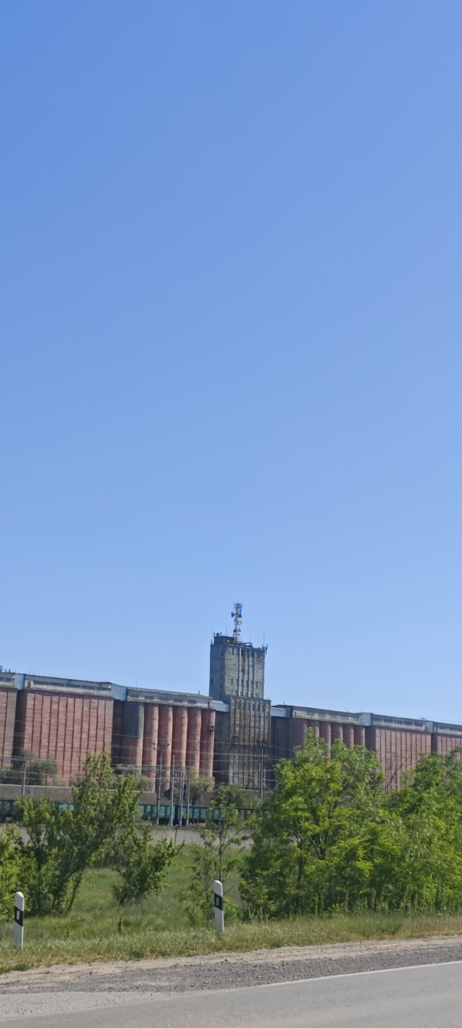 The image shows a large grain elevator complex in Dubovskoye, Russia, under a clear, bright blue sky. The buildings are predominantly red and gray, consisting of long, horizontal structures with cylindrical silos. A central tower rises above the complex, topped with communication antennas. Green trees and foliage are visible at the base of the structure, adjacent to a road marked with guard posts.