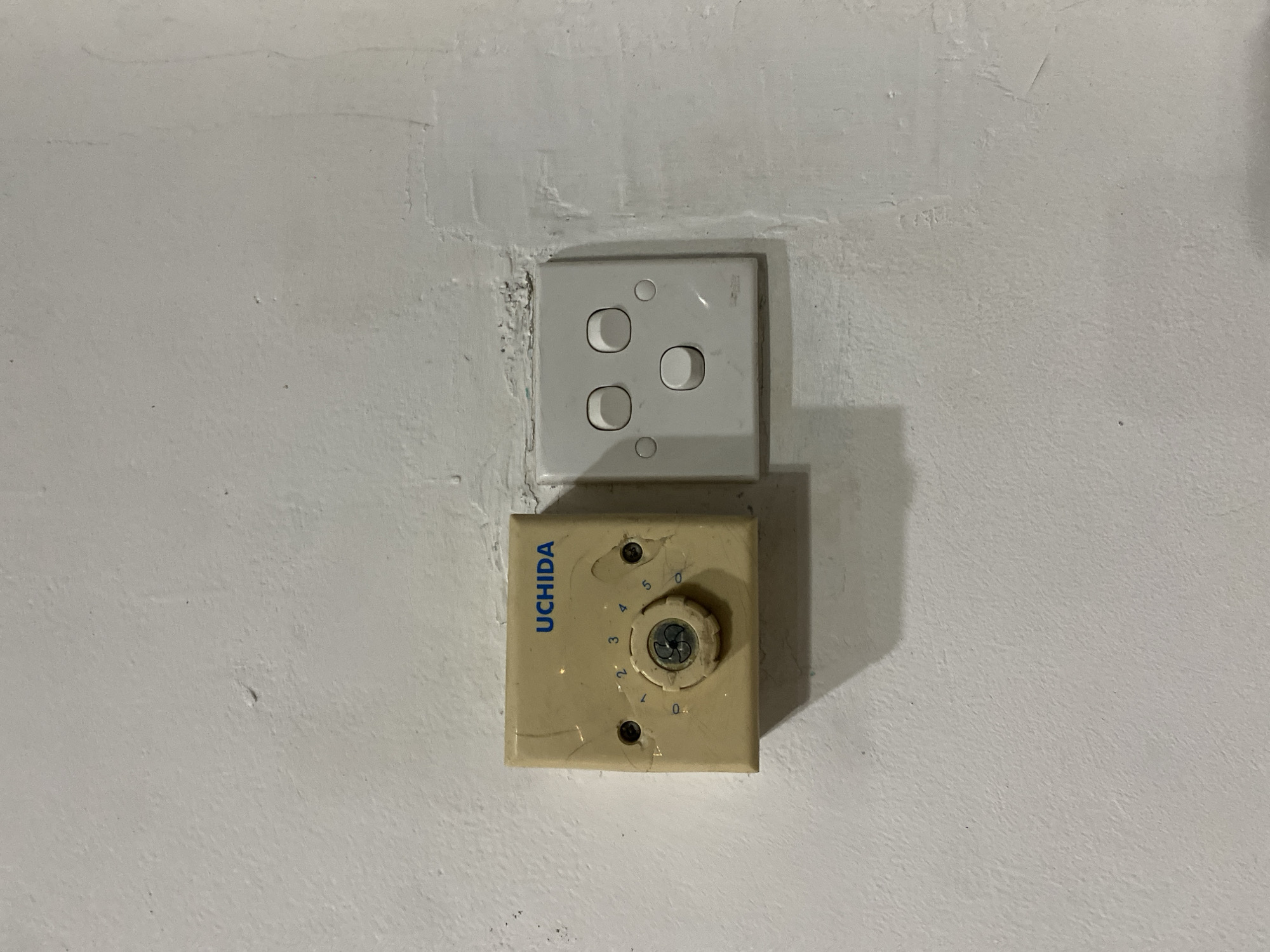In Kuta Selatan, Indonesia, a close-up shot captures an electrical outlet and a vintage rotary dial switch mounted on a white, textured wall. The rotary switch, cream-colored and square, features the "UCHIDA" brand name in blue lettering. A circular dial with numbers ranging from 0 to 5 is visible, with a central screw and a smaller dial in the center. To the right, a standard white electrical outlet with three rocker switches is present. The wall exhibits minor imperfections, including scuffs and textured patches, suggesting an indoor environment. No people or specific activities are depicted.