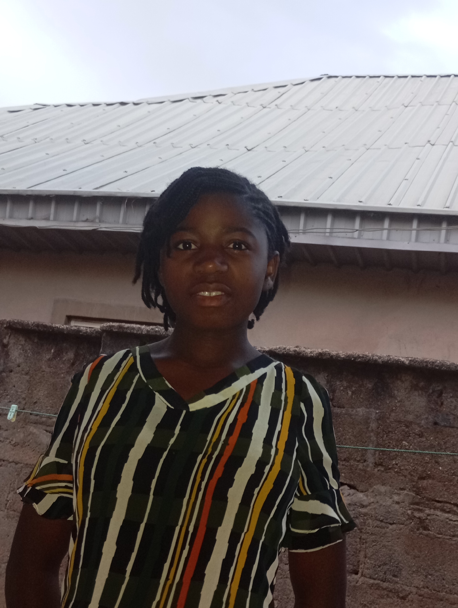 A portrait-style photograph captures a young Black individual, likely female, standing outdoors in what appears to be a residential setting in Jos, Nigeria. The person is positioned centrally, looking directly at the camera with a neutral to slightly alert expression, her mouth slightly open to reveal some teeth. She has dark skin and her hair is styled in braids, pulled back from her face.
She is wearing a V-neck, short-sleeved top featuring a bold pattern of vertical stripes in various widths and colors, including dark green, off-white, orange, and yellow.
The background features a light-colored, corrugated metal roof of a building, stretching across the upper half of the frame under an overcast sky. Below the roof and behind the subject, a light brown wall is visible. In the lower background, a textured, dark brown concrete wall or fence runs horizontally. A thin clothesline, with a white clothespin attached, is visible on the left side, strung in front of the concrete wall. The soft, even lighting suggests either an overcast day or the transition hours of early morning or late afternoon. No text is visible in the image.
