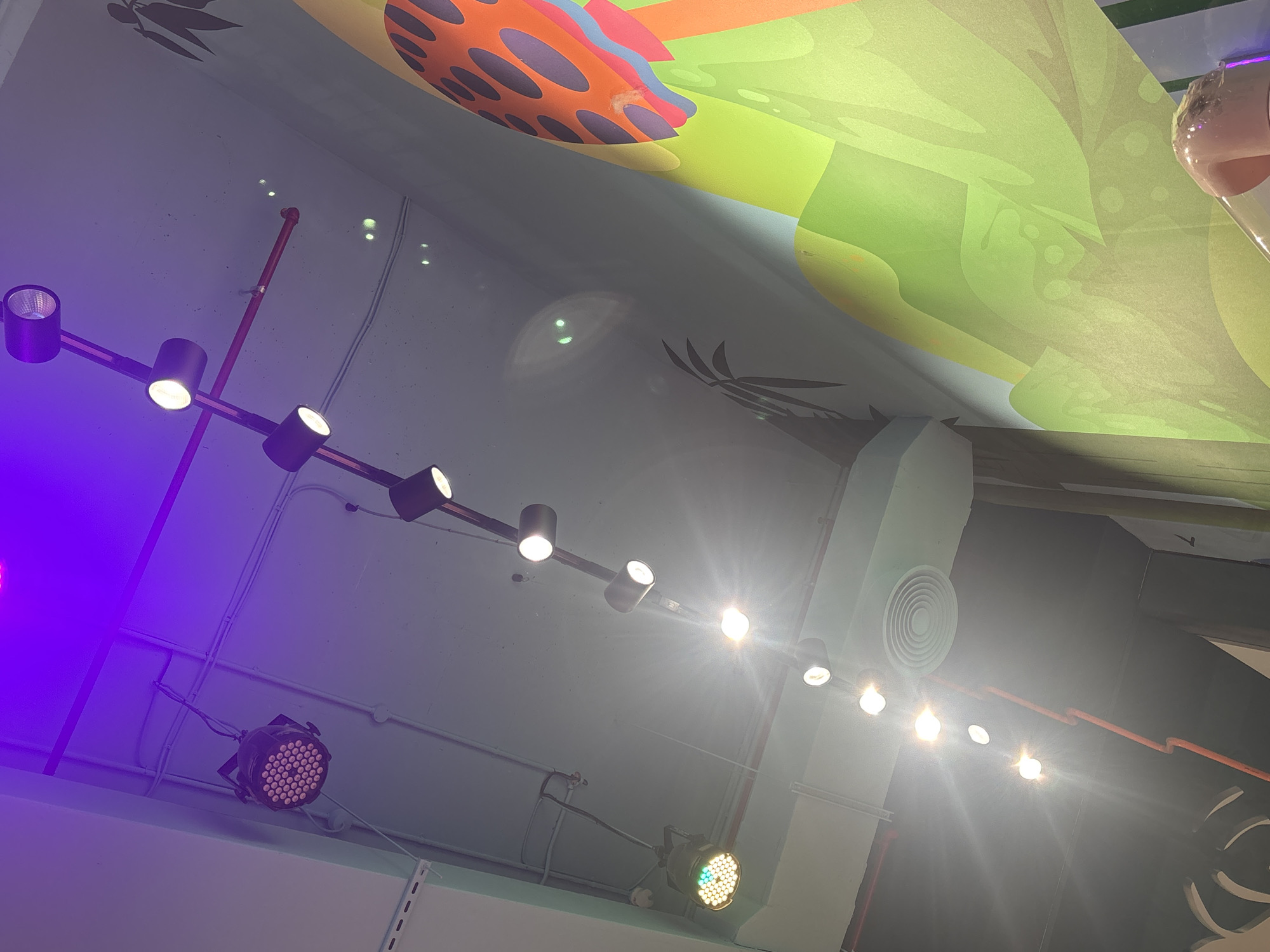 The image shows a ceiling with various lights and a mural. There are several track lights with cylindrical lamps, some emitting bright white light and others appear to be purple or blue. One light fixture is a black box with many small circular bulbs. There are also red pipes and conduits visible along the ceiling. A mural is painted on the wall, featuring abstract shapes in bright green, orange, and purple, resembling a cartoonish landscape with elements that could be interpreted as hills or mushrooms. The overall impression is that of an indoor recreational space, possibly a play area or entertainment venue, illuminated by a combination of ambient and focused lighting. The image is taken from a low angle, looking upwards towards the ceiling and the mural.