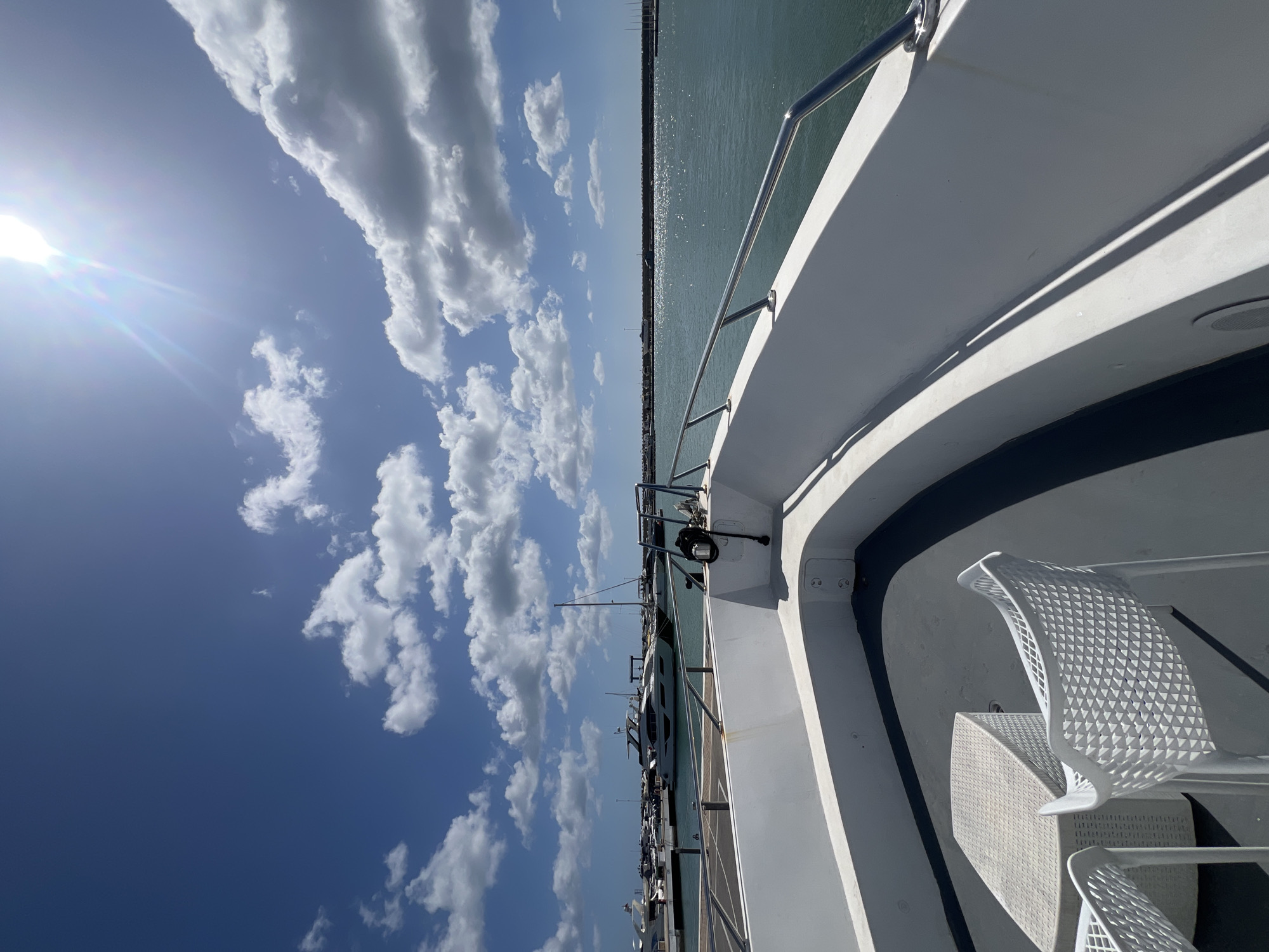 The image, taken in M'Diq, Morocco, presents a scene from the deck of a boat or yacht. The view includes the sky, filled with fluffy white clouds against a blue backdrop, and the sun emitting bright rays. The water is visible, reflecting sunlight. The foreground of the image features a portion of the boat's structure, including the white exterior, a railing, and a white, modern-looking chair and table set. Other boats are visible in the distance.