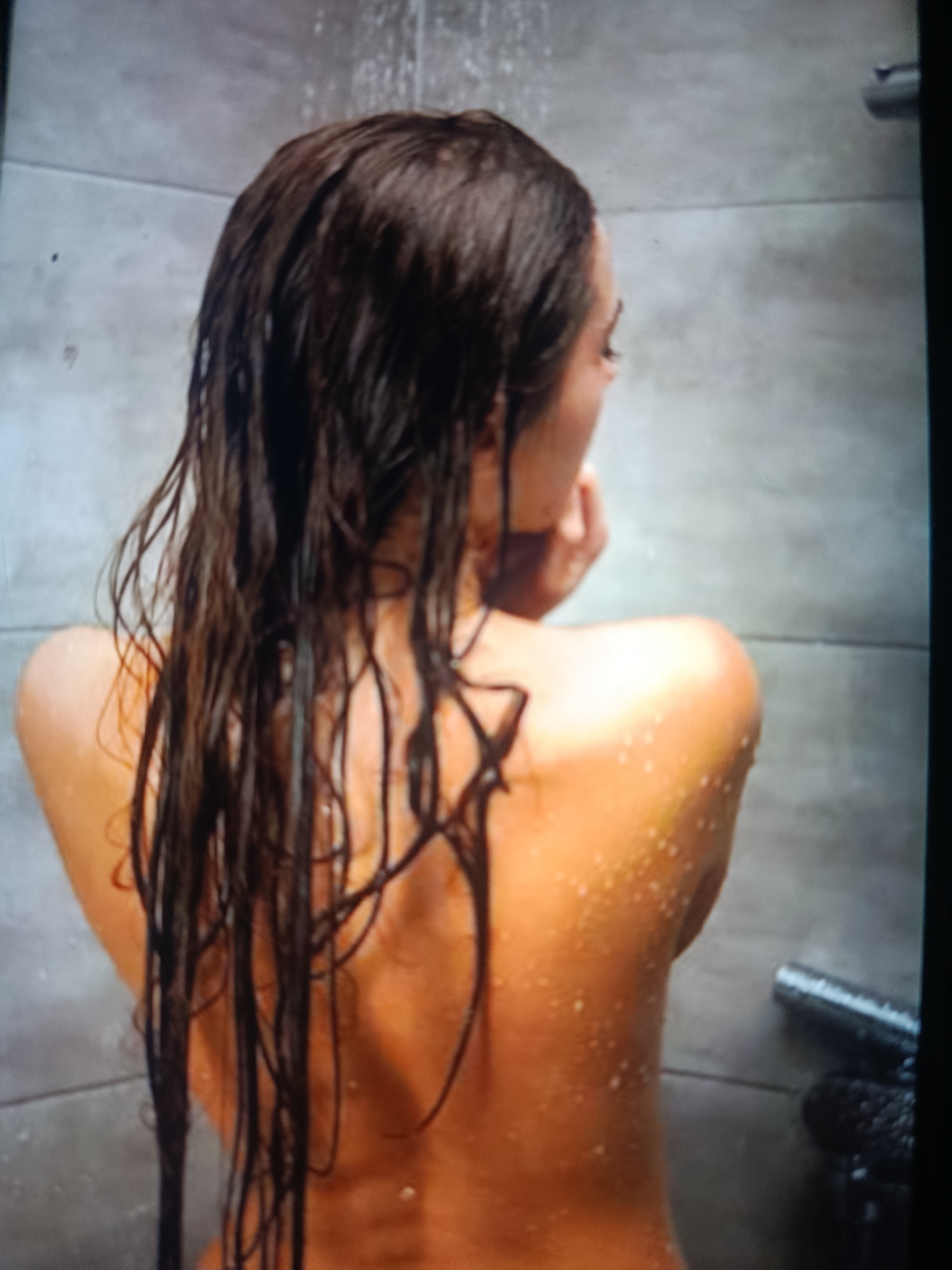 A woman with wet, dark brown hair is standing in a shower. The shower has gray tiles and a silver shower head. Water is running down her back and shoulders. Her back is bare and covered in water droplets. The location in the city of Juan Griego, Venezuela, is not evident in the image.
