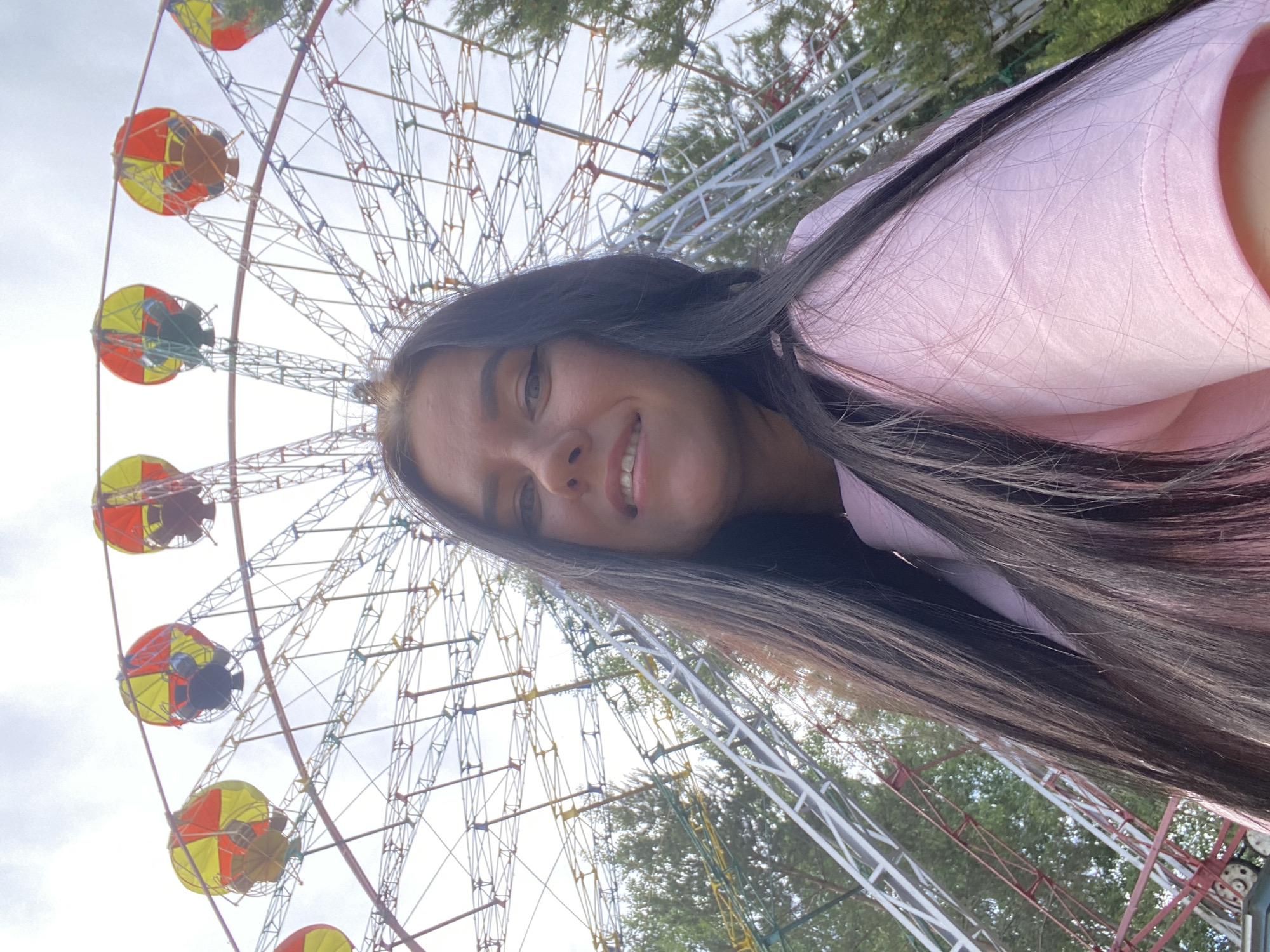 A selfie-style image, rotated ninety degrees counter-clockwise, depicts a young woman with long, dark hair smiling happily. She is wearing a light pink top, and her face is positioned in the upper-right portion of the frame. Her dark hair sweeps across her shoulder and the pink fabric.
In the background, a large Ferris wheel with an intricate metal framework dominates the scene. The Ferris wheel features multiple colorful gondolas, distinctively painted in yellow and red segments. The structure of the ride is set against a bright, overcast sky, indicating daytime. Scattered green trees and foliage are visible through the gaps in the Ferris wheel's structure, suggesting an outdoor, park-like environment.
The overall scene conveys a cheerful atmosphere, likely at an amusement park or fairground in Chaikovsky, Russia. The woman's expression suggests enjoyment. There is no visible text in the image.