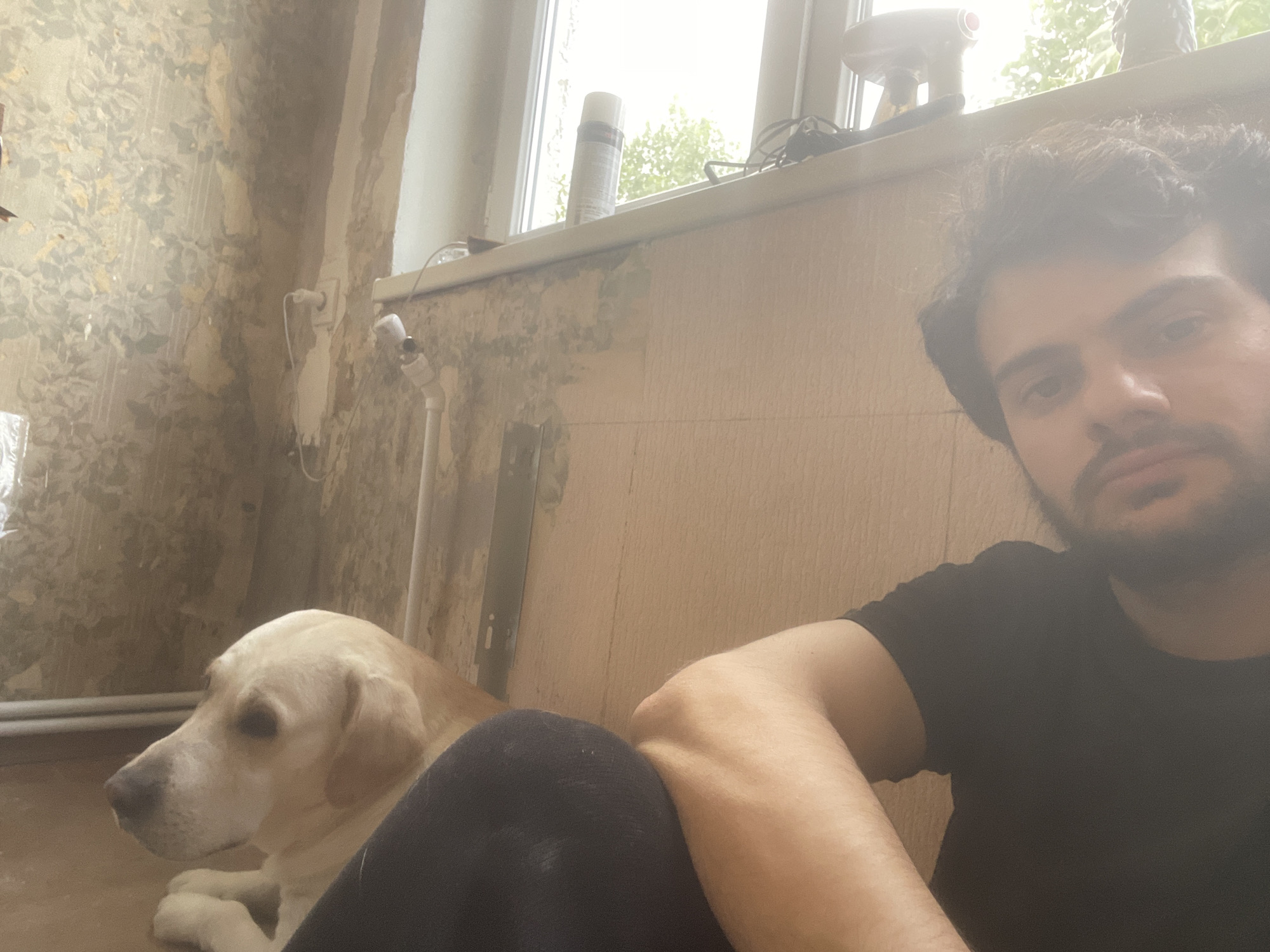 A light-colored Labrador retriever is lying on the floor next to a person wearing a black t-shirt. The dog is looking to its left. The person is in the foreground and appears to be looking at the camera. The background shows a window with some items on the sill, including a can of spray and a hairdryer. The walls have peeling wallpaper and visible pipes. The overall impression is an indoor, domestic setting, likely a residential space. There are no identifying markers that would suggest a specific location within Tbilisi, Georgia.