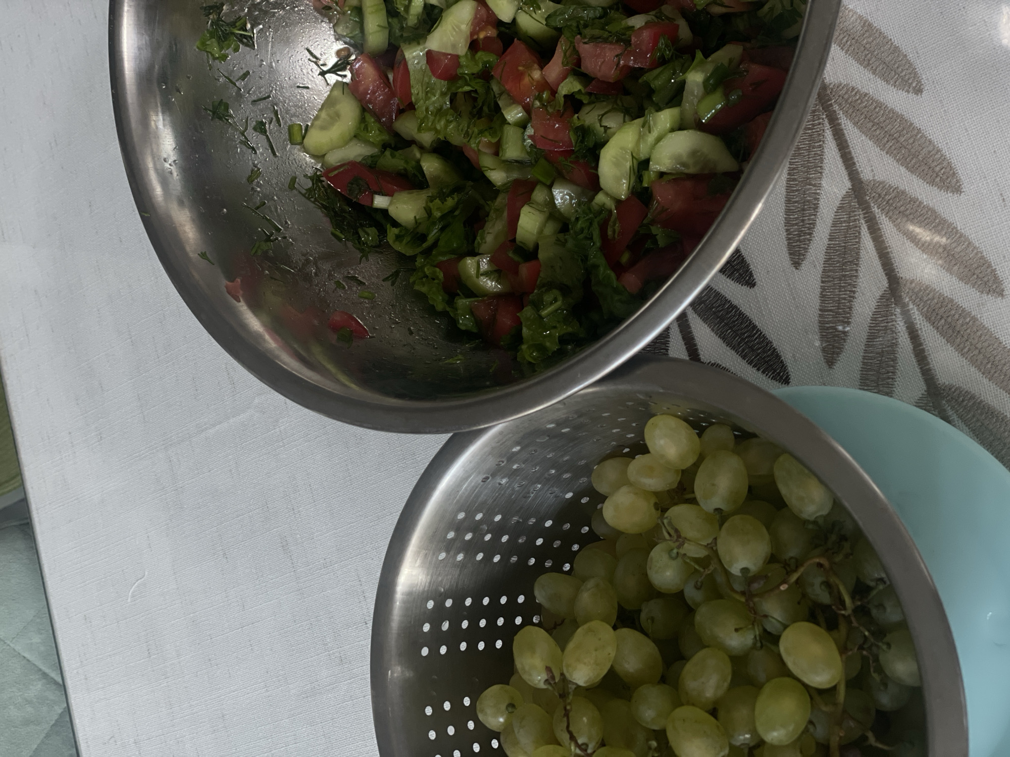 A close-up, high-angle shot shows a stainless steel colander filled with green grapes, placed next to a large stainless steel bowl containing a chopped salad of tomatoes, cucumbers, and herbs. The colander is partially resting on a light blue dish. The objects are situated on a white tablecloth with a subtle pattern of brown leaves. The background features a light green textured surface. The scene is indoors, and the lighting suggests it is daytime. No people are visible. The location is identified as Angarskie Khutora, Russia.