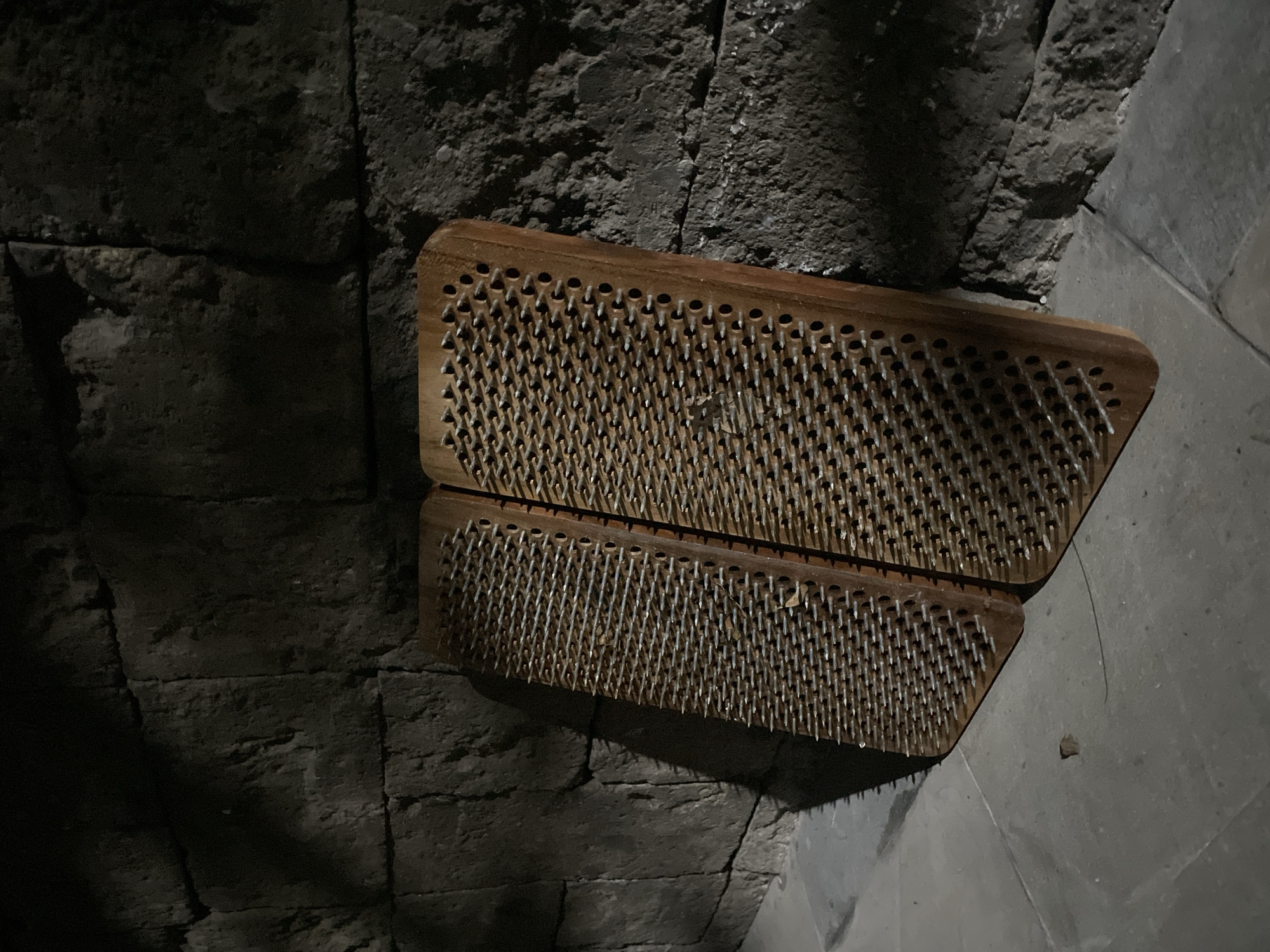 Two wooden Sadhu boards, or nail boards, are the central subjects of this dim, static image. Each board is rectangular, crafted from light brown wood, and densely embedded with numerous sharp, silver-colored metal nails. The nails are arranged in parallel rows, pointing upwards, forming a surface often associated with meditative practices or endurance challenges. The two boards are positioned slightly offset from each other, leaning against a rough, dark gray wall in what appears to be a corner.
The setting consists of a rugged, textured wall, likely made of stone or coarse concrete, exhibiting prominent seams and an uneven surface. This wall forms the background and extends to the left, characterized by deep shadows. To the right and beneath the boards, the surface transitions to a smoother, lighter gray concrete, suggesting a floor or an adjacent wall within an enclosed space.
The lighting is low and uneven, creating strong contrasts and pronounced shadows cast by the nail boards onto the wall and floor. This dramatic illumination contributes to a stark and quiet atmosphere. There are no people visible, and no activity is taking place. The image evokes a sense of stillness and discipline, consistent with the functional purpose of the objects. No text is visible. The location context is Kuta Selatan, Indonesia.