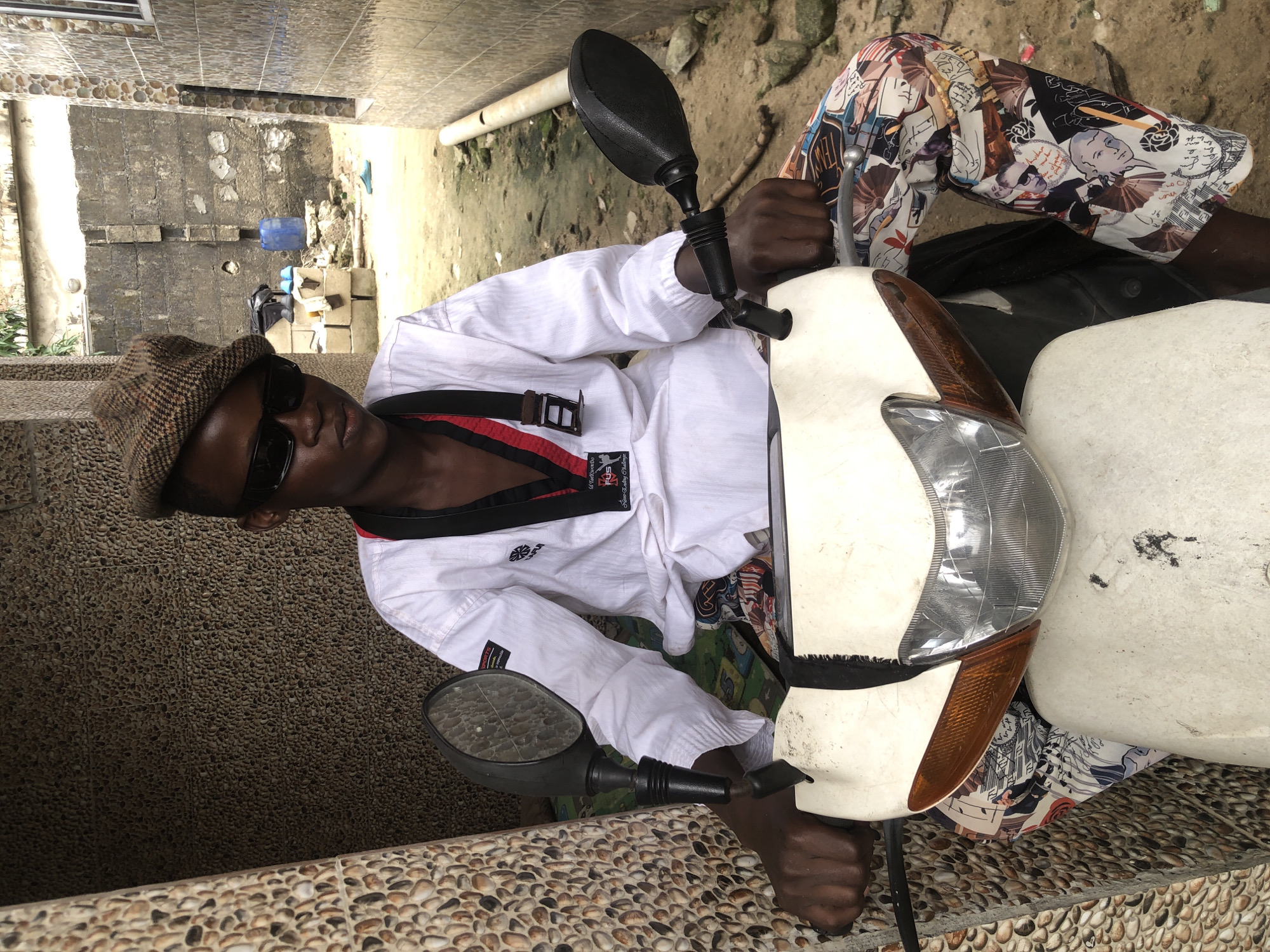 A young person wearing a white taekwondo uniform, black sunglasses, and a checkered cap sits on a white scooter in Ajah, Nigeria. The individual's legs are clad in colorful patterned pants featuring various illustrations. The scooter's front is visible, with a headlight and orange turn signal. The background depicts a narrow outdoor area between buildings, with a textured wall on the left, a concrete wall with some debris on the right, and a dirt path.