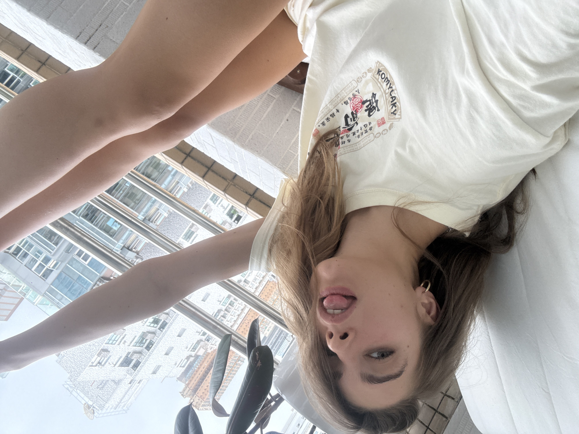 A woman is lying on a white surface, possibly a bed or couch, in what appears to be an upside-down view. She is wearing a light-colored T-shirt with text and imagery in what seems to be Asian characters and English words "KOMYLAKY" printed on the chest. The woman has medium-length brown hair, and is sticking her tongue out. She is wearing a gold hoop earring in her left ear. A plant is partially visible in the lower left corner of the image. Through the window behind her, there's a view of tall buildings, suggesting a city environment, possibly Hong Kong, China.