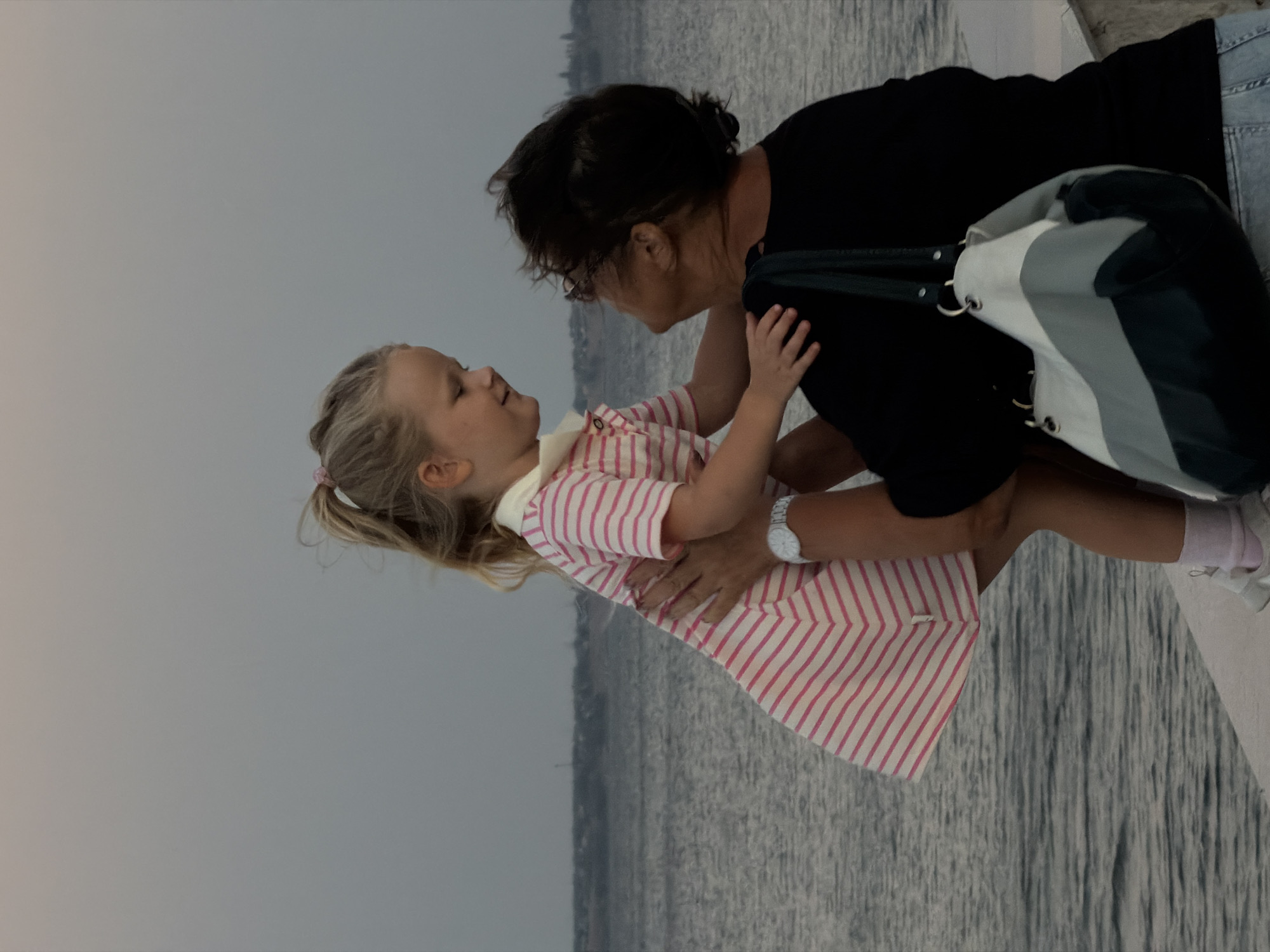 An outdoor, low-angle, medium shot captures a woman holding a young blonde girl up in the air by the sea at dusk in Kamyshin, Russia. The girl, wearing a pink and white striped dress and pink socks, smiles and looks down at the woman. The woman, wearing a black t-shirt and glasses, looks up at the girl. She has a multi-colored bag with her. The water in the background is dark and choppy, with a faint horizon line and some distant structures. The sky is overcast and a pale grey.