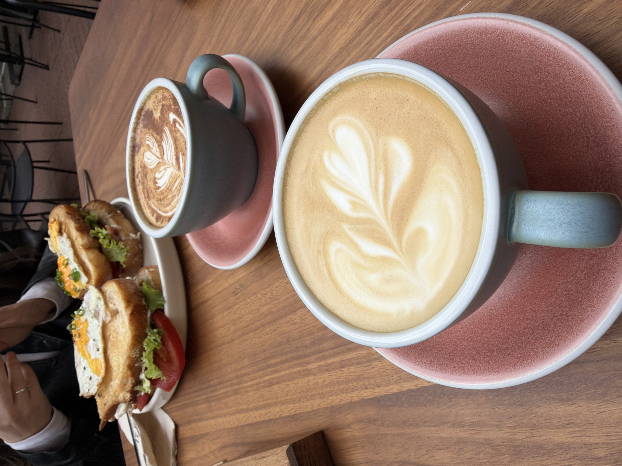 On a wooden table, there are two coffee cups with latte art and a plate of sandwiches. The larger coffee cup, filled with a lighter-colored latte, has a white rim and is sitting on a pink saucer. Its handle is light blue. Beside it, there's a smaller coffee cup containing a darker latte on another pink saucer. A plate with two sandwiches, filled with eggs, tomatoes, and lettuce, is placed near the smaller cup. A person's hand and part of a black jacket are visible on the left. The exact location in Sofia, Bulgaria, cannot be determined from the image alone.