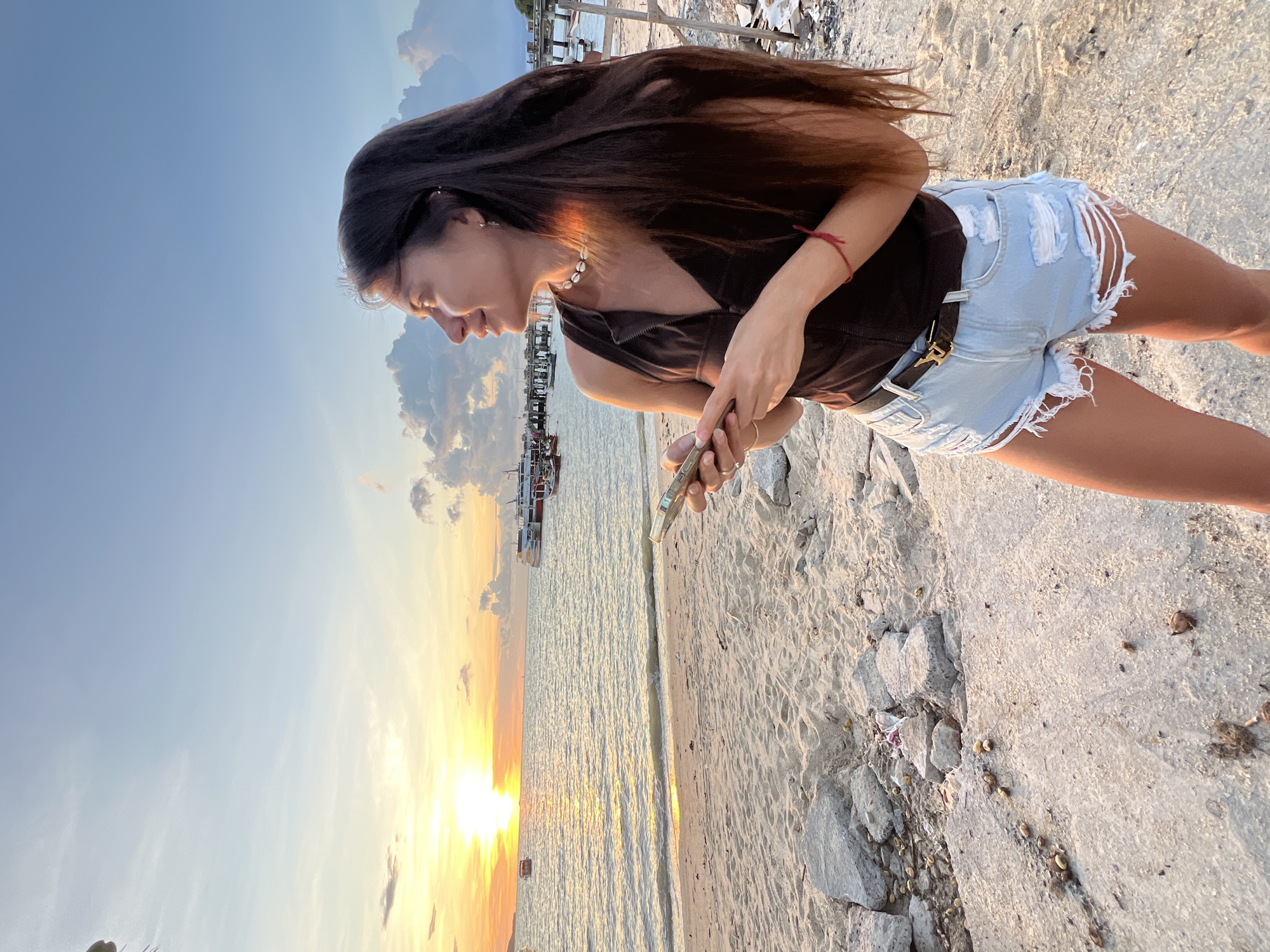 A woman stands on a sandy beach in Baan Bang Rak, Thailand, holding a smartphone. The sun is setting over the ocean, casting a warm glow on the scene. The woman is wearing a brown tank top and ripped denim shorts. In the background, several boats are docked near a pier. The beach is littered with rocks and debris.