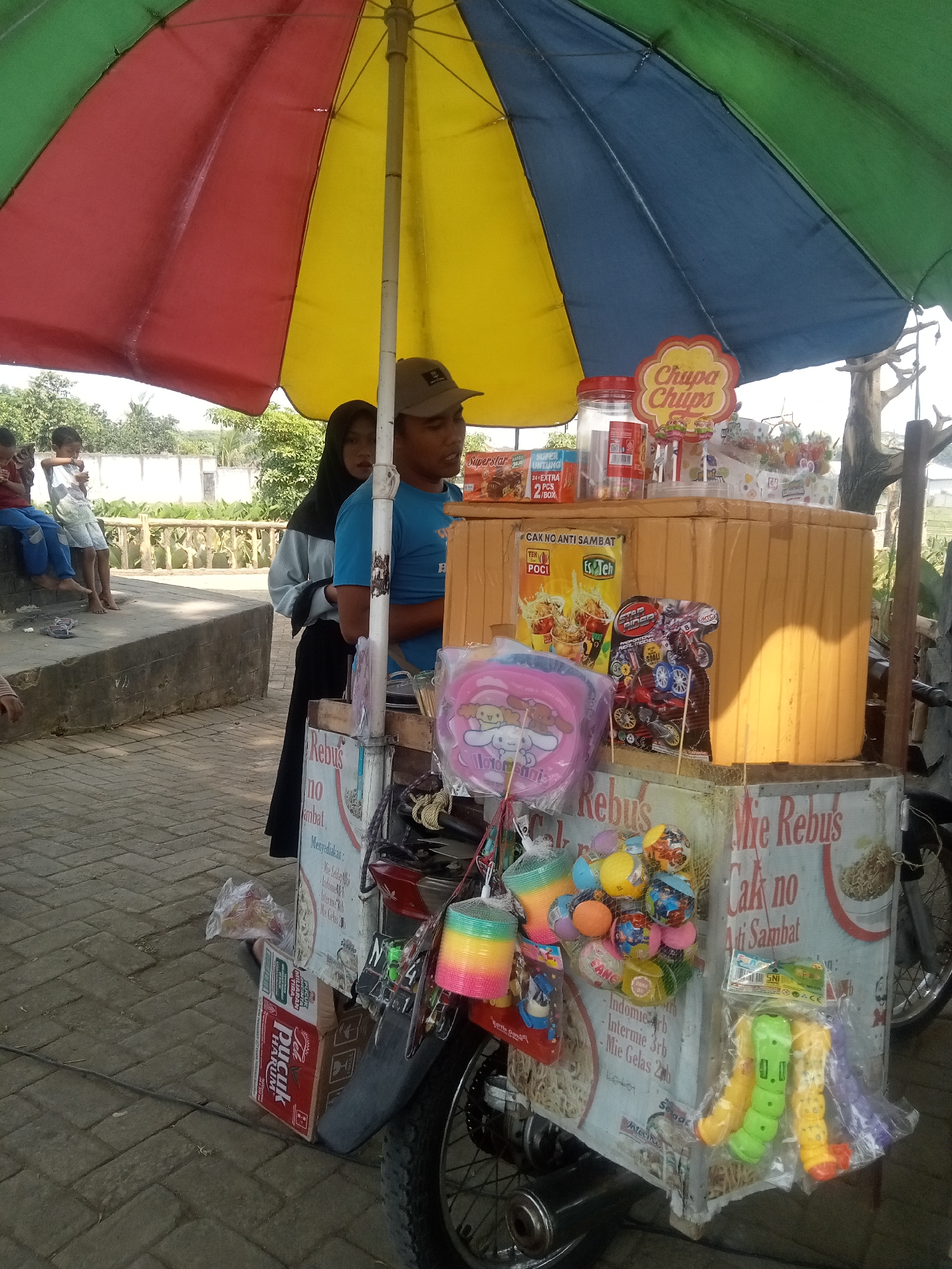 In Malang, Indonesia, a street vendor sells snacks and toys from a cart beneath a large, multicolored umbrella. The vendor, a man wearing a blue t-shirt and a cap, stands behind the cart. Beside him, a woman in a hijab and dark clothing observes. In the background, children are visible, seated on a concrete ledge. The cart is stocked with various items, including bags of snacks, candies, small toys, and what appears to be instant noodles. A motorcycle forms the base of the cart, its wheel visible on the right. The ground is paved with interlocking bricks. The scene is set outdoors, with foliage and structures in the distance, indicating an urban or suburban environment.