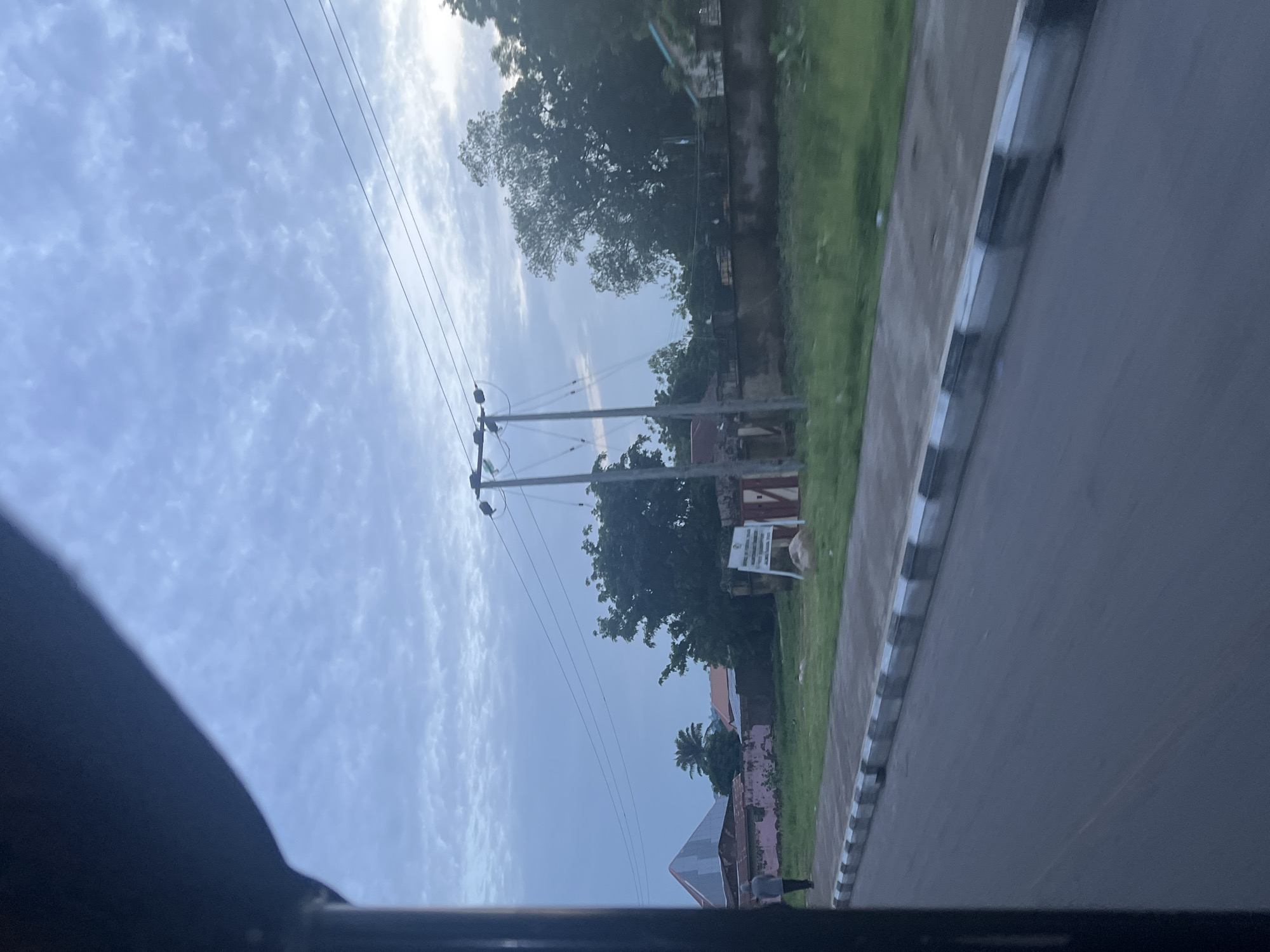The image captures a view from a moving vehicle in Jalingo, Nigeria, likely during early morning or late afternoon given the soft light and cloud cover. In the foreground, a dark, curved structure, possibly the interior of the vehicle, frames the left side of the scene. The road surface is visible, with a distinct curb marked with alternating black and white stripes.
Beyond the curb, a paved walkway runs alongside a grassy verge. A single pedestrian, seen from the back, walks away from the camera on the walkway. Their attire is casual, a light-colored shirt and dark pants.
The middle ground of the image reveals a residential or institutional area. Several buildings with varied rooflines and colors are present, including one with a distinctive pyramidal roof. A tall palm tree stands among other foliage. A white signpost with text is visible, positioned on the grass. Two utility poles with electrical wires strung between them stand prominently.
The background is dominated by a serene sky filled with light, scattered clouds. Lush green trees and a concrete wall are also part of the scene, suggesting a well-established area within the city. The overall impression is one of a typical urban streetscape in Jalingo.