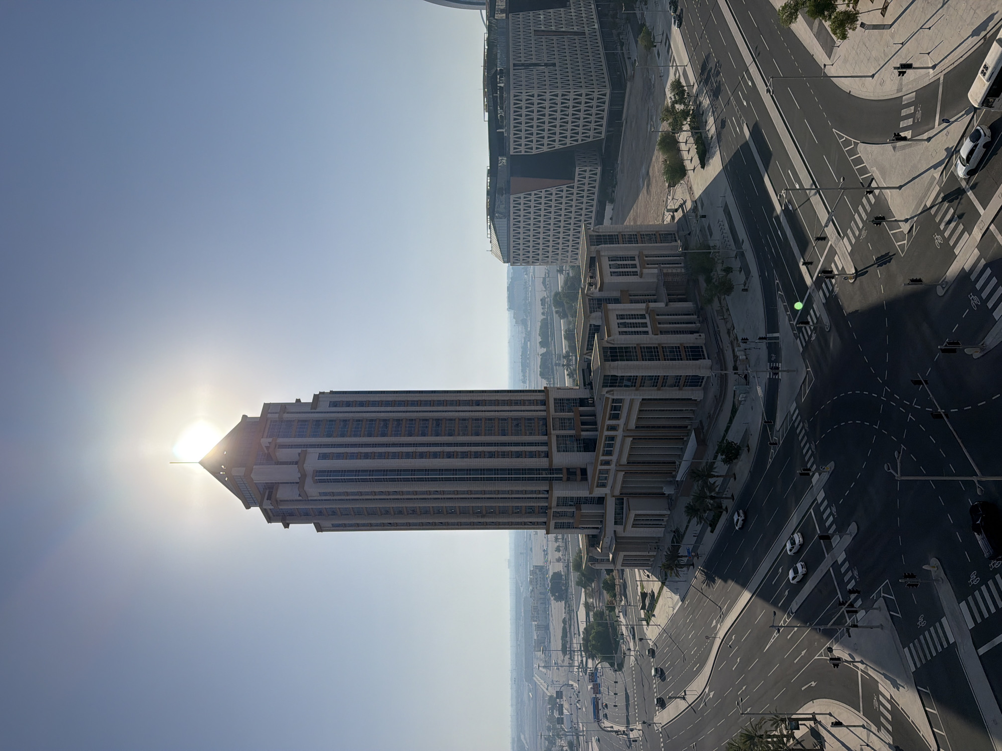 This media file depicts an urban landscape from a high vantage point, likely a building. The sun is low in the sky, casting long shadows and creating a bright glare that partially obscures a tall, modern office building in the foreground. The building is characterized by its rectangular shape, numerous windows, and a peaked roof. Adjacent to this building, another, larger structure is visible, featuring a distinctive patterned facade with a triangular motif.
The scene also includes a complex network of roads with multiple lanes, traffic lights, and pedestrian crossings. Several cars, including white sedans and a white bus, are navigating the intersections. Palm trees are scattered along the roadways, suggesting a warm climate. The overall impression is one of a contemporary city, possibly in the Middle East, given the architectural style and vegetation. The time of day appears to be either early morning or late afternoon due to the low angle of the sun and the resulting shadows. The sky is clear and a pale blue, indicating fair weather. No people are distinctly visible, and there is no visible text within the image.