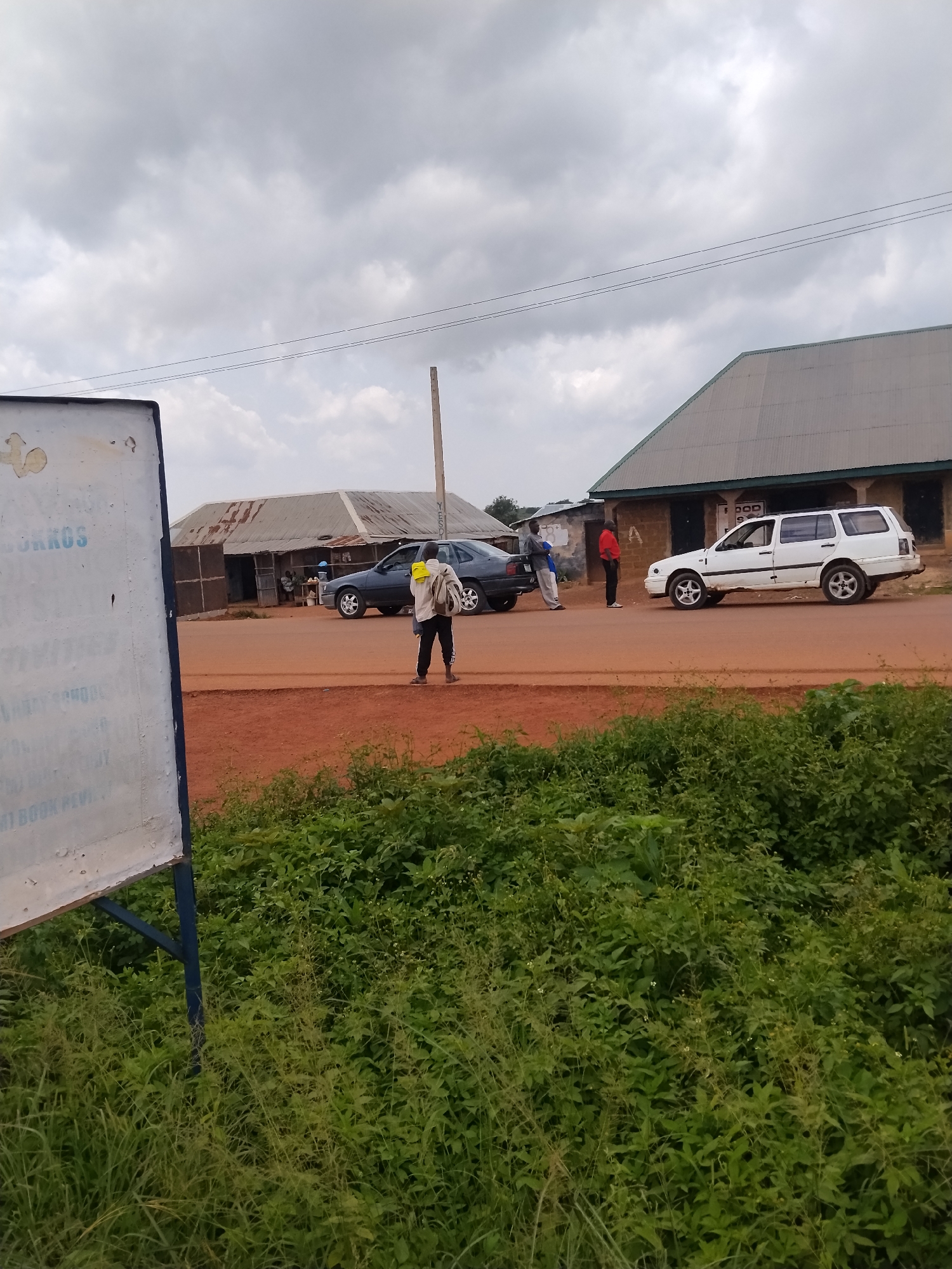 In the city of Durumi, Nigeria, an outdoor scene unfolds on a dirt road under a cloudy sky. A white van and a dark sedan are parked on the side of the road. A man in a red shirt stands near the white van, and another man in a grey shirt and white pants stands near the dark sedan. A third man, wearing a light-colored backpack, walks away from the camera, holding a yellow object. In the foreground, a portion of a white sign with blue lettering is visible, and dense green foliage lines the bottom of the frame. A tall, thin pole stands to the right of the vehicles, and utility lines are strung across the sky. Buildings with corrugated metal roofs are visible in the background, suggesting a commercial or residential area. The ground is covered in reddish-brown dirt.