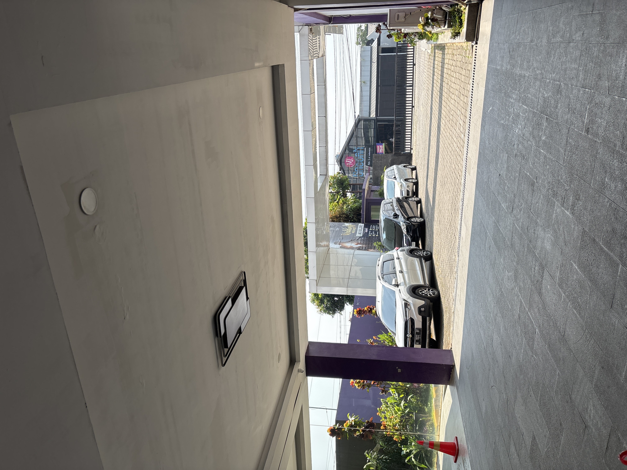 The image shows an outdoor scene in front of a building with a purple column in the foreground. A white SUV is parked on the right, with its license plate reading "A 1022 BY". To the left of the SUV, a black car is parked. Further into the background, there is a building with a sign that says "ML Tanconff". The area appears to be a parking lot or driveway. The sky is visible in the background, and there are trees and plants on the left side of the image. The ground is paved with grey tiles. The image was taken in Binong, Indonesia.